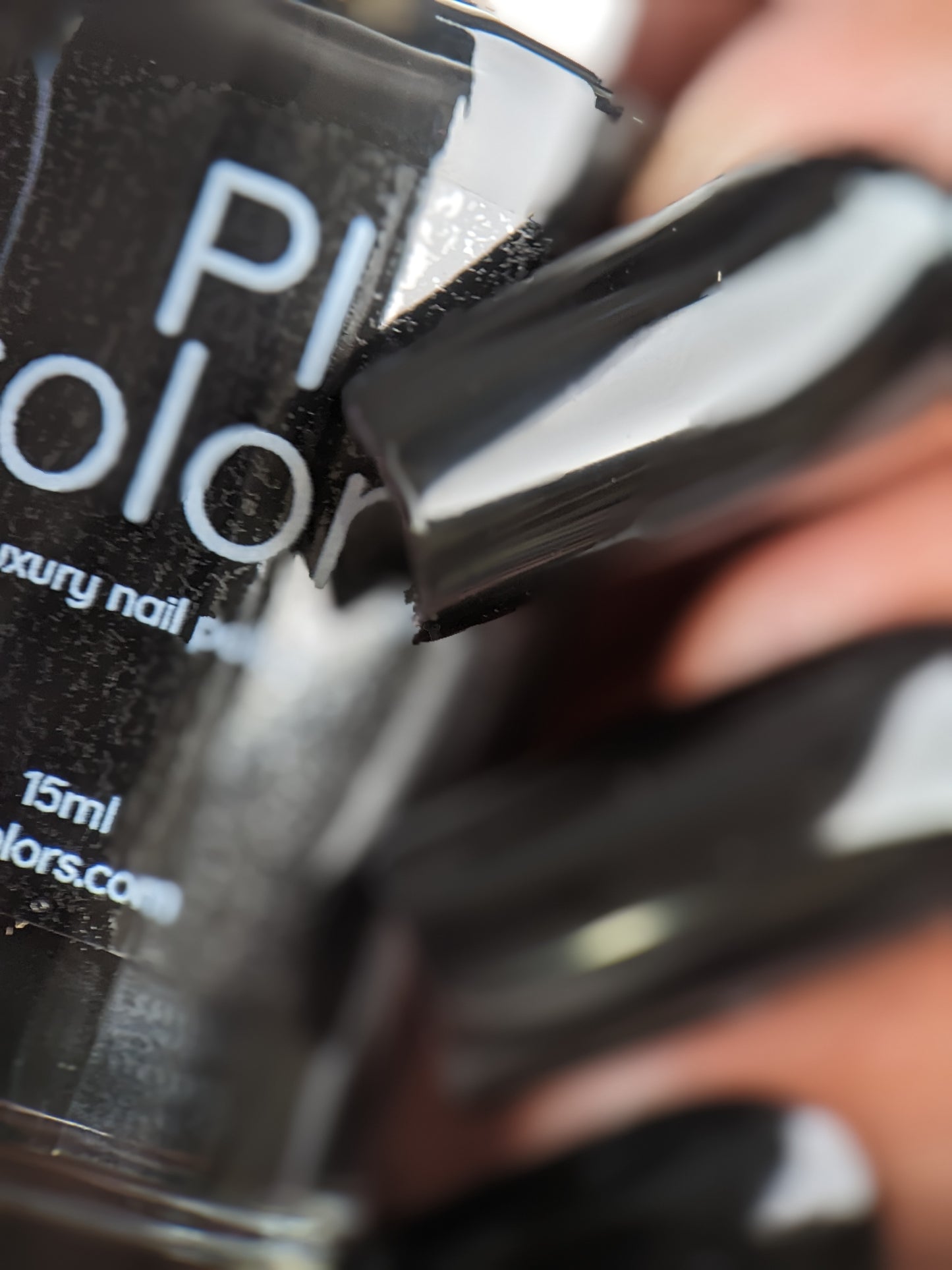 Close-up of a hand with long, glossy black fingernails holding a bottle of PI Colors' Onyx.274 Black Nail Polish. The partially visible text on the bottle indicates it contains 15ml of polish and highlights the shiny creme finish of the nails.