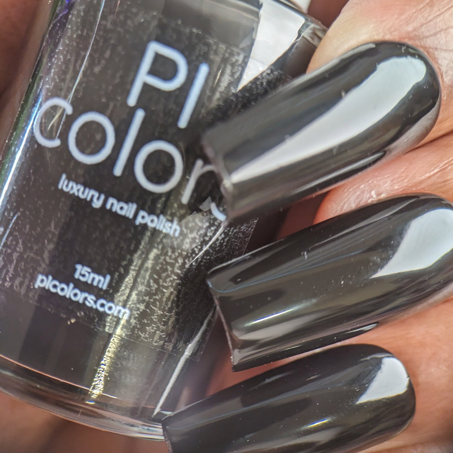 Close-up of a hand with long nails adorned in glossy dark gray, nearly black nail polish, holding a bottle of Onyx.274 Black Nail Polish by PI Colors. The label on the bottle reads "15ml, pi.colors.com," highlighting its 10-free formula and glossy crème finish.