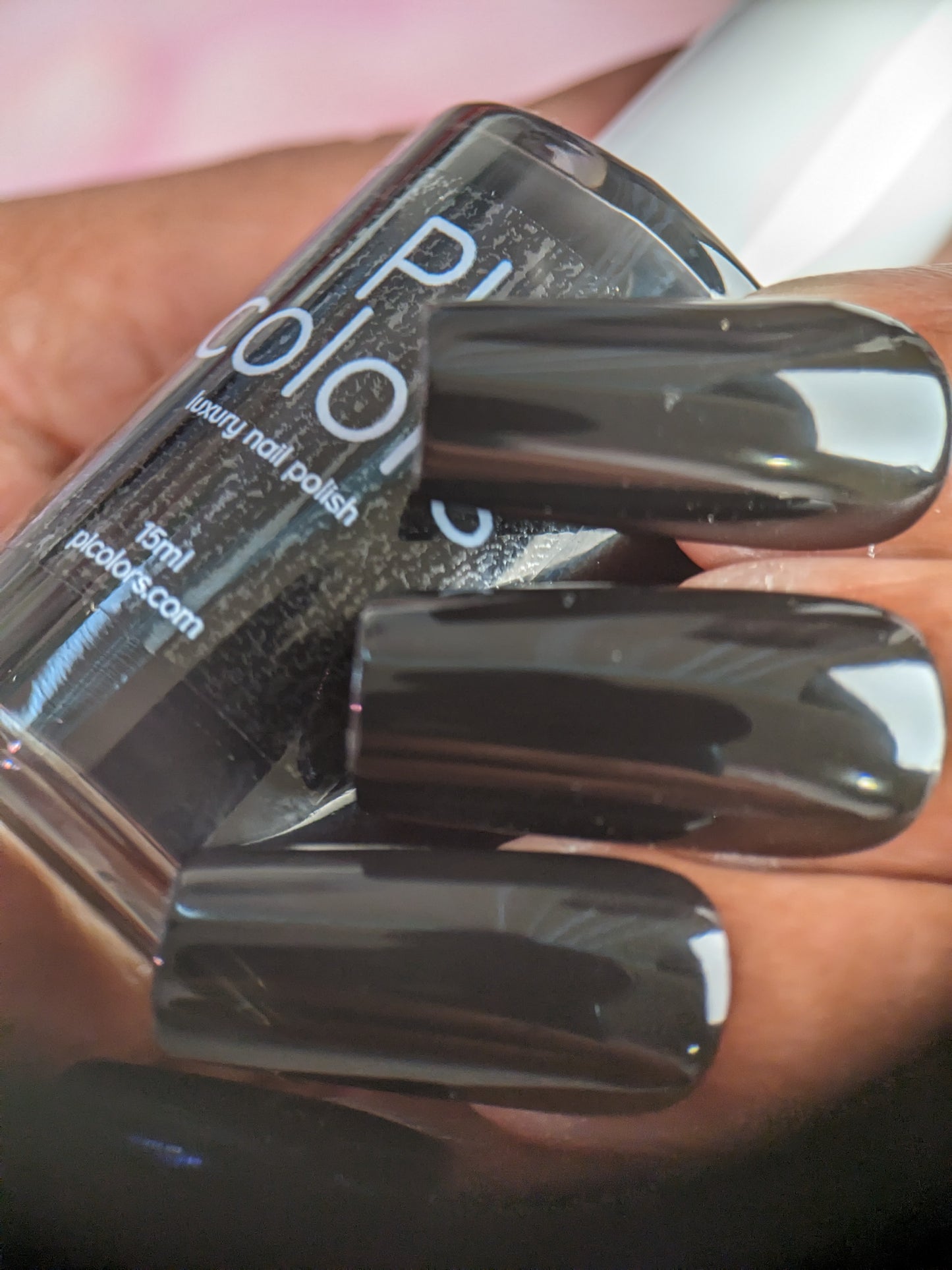 A close-up of a hand with long nails painted in PI Colors' Onyx.274 Black Nail Polish is showcased. The hand holds a "Pure Color" bottle labeled as "15ml," featuring an exquisite glossy creme finish. This 10-free black nail polish ensures a chic and polished look without harmful chemicals.