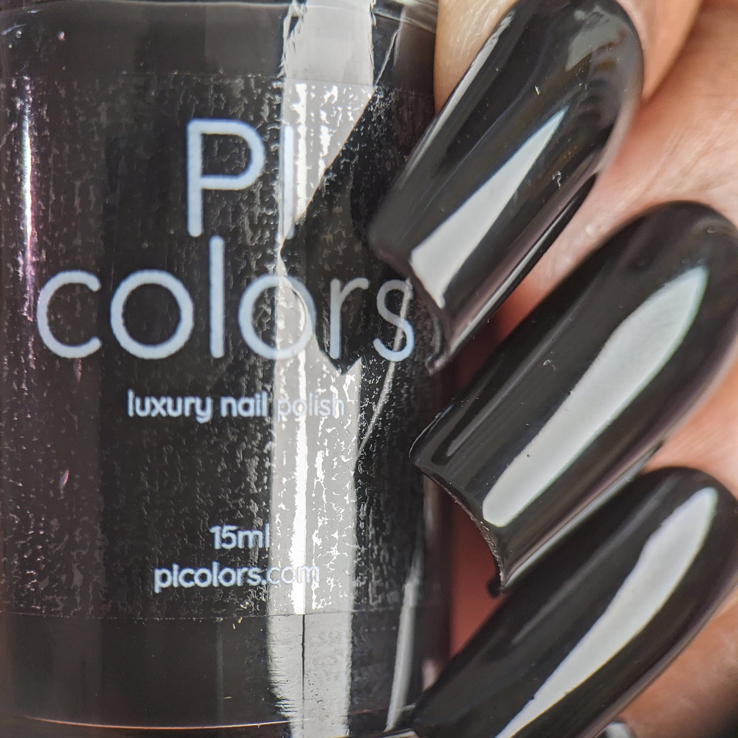 A close-up of a hand with long, glossy black nails in a creme finish is holding a bottle labeled "Onyx.274 Black Nail Polish" by PI Colors. The background features a textured, reflective surface.