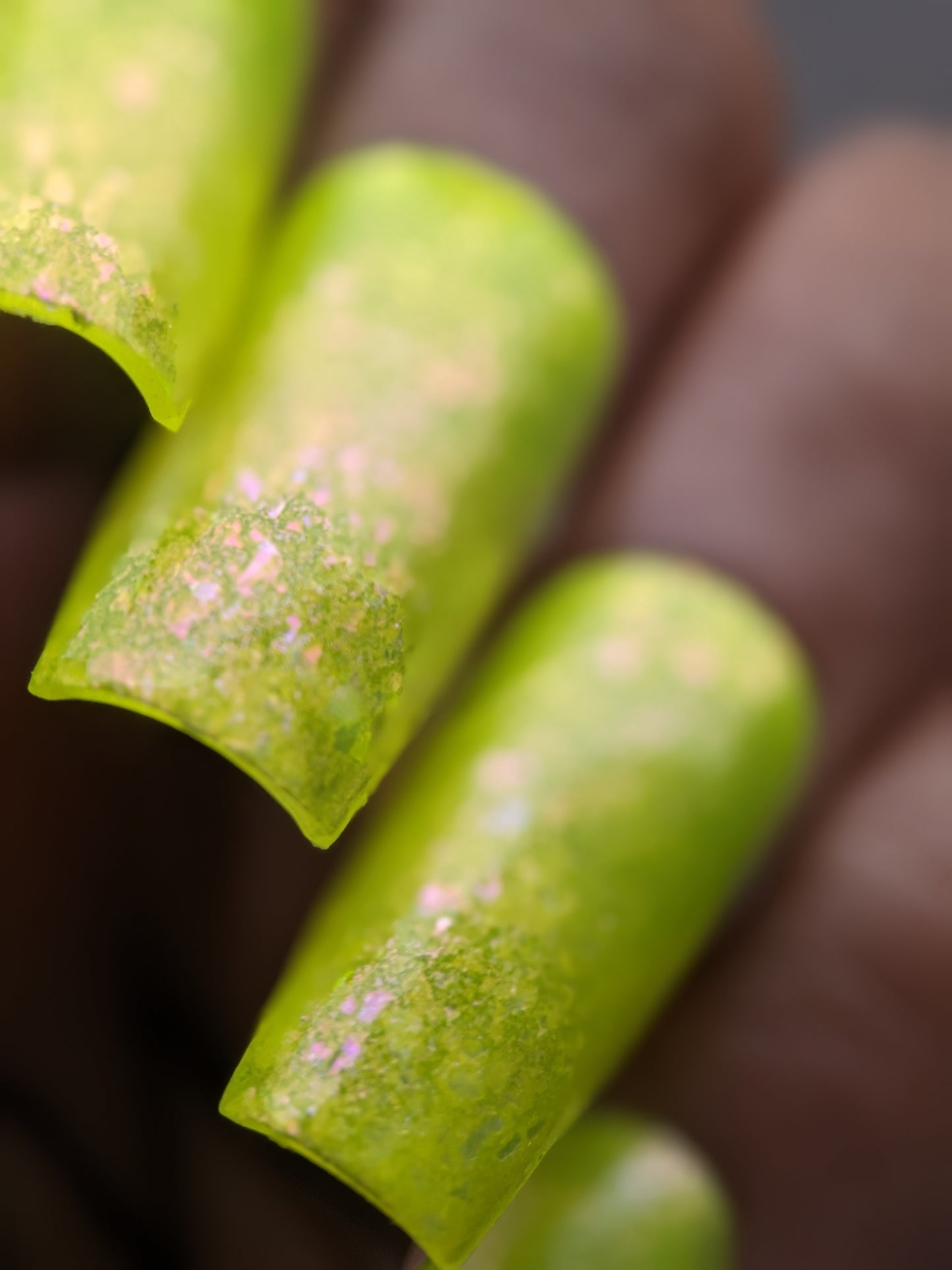 Close-up of nails adorned with PI Colors' Neon Shine Yellow, a luxury nail lacquer in a bright yellow-green shade with an iridescent finish. The handmade polish provides a textured appearance, while the nails are rounded and slightly squared. The background is blurred and neutral.