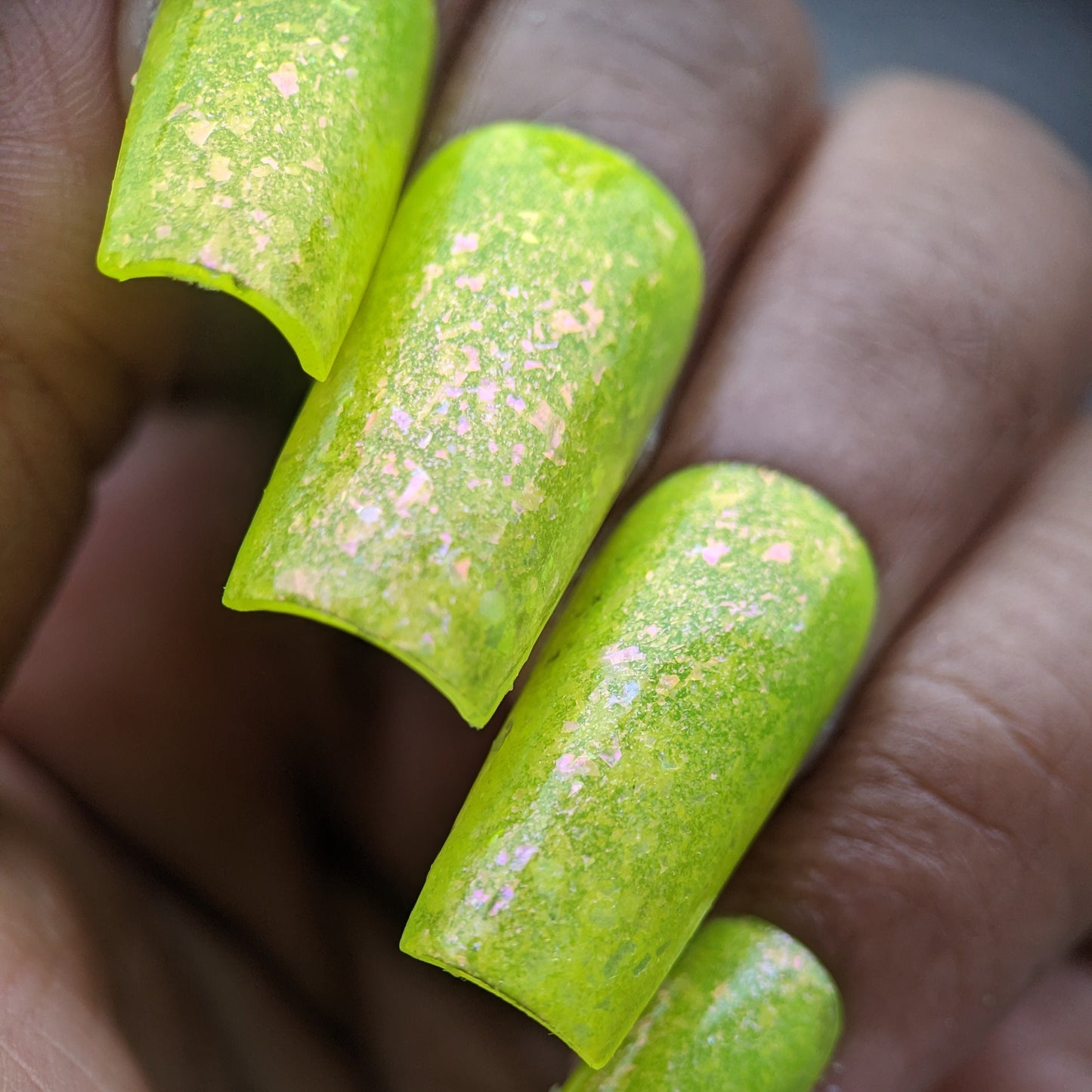 A close-up of a hand with long, coffin-shaped nails highlights the vibrant Neon Shine Yellow polish by PI Colors. This handmade polish features iridescent flakes that create a shimmering effect under the light, offering an elegant finish akin to luxury nail lacquers.