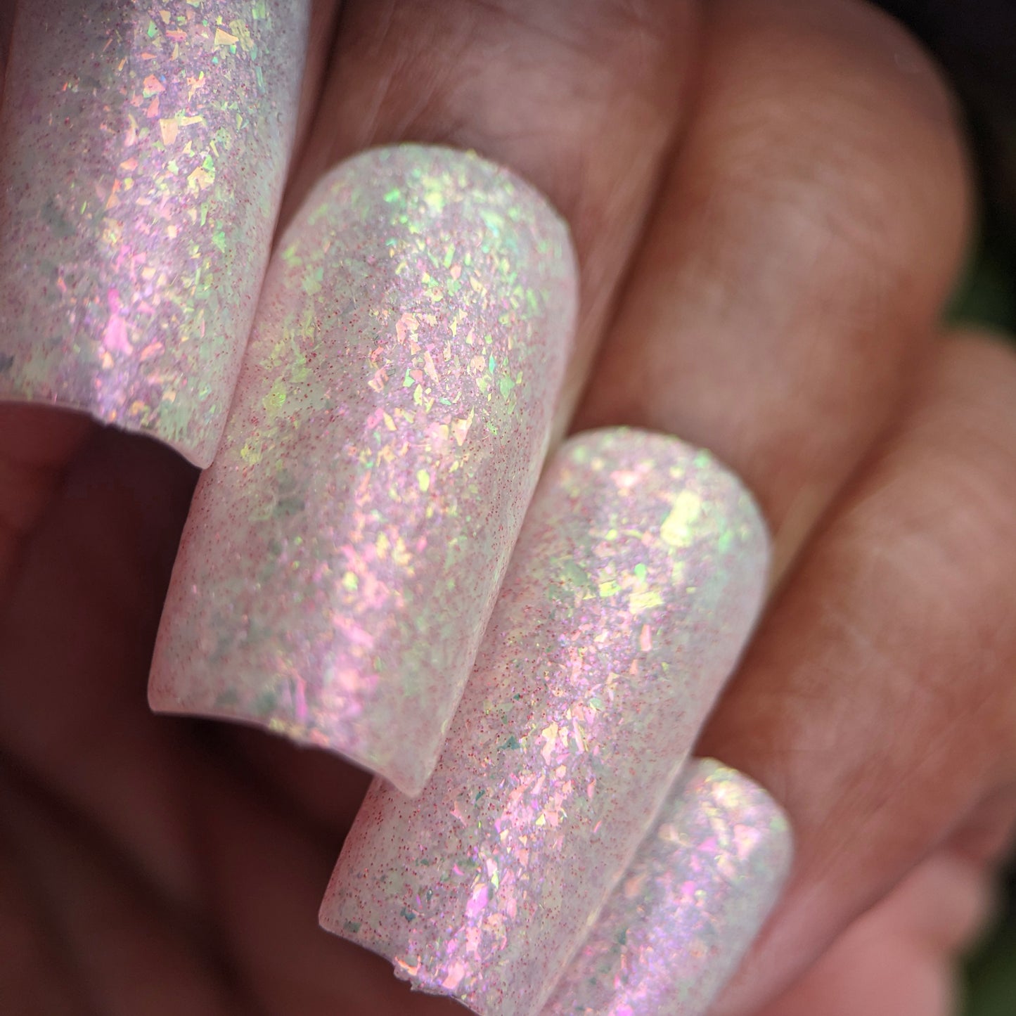 A hand with nails painted in PI Colors' Caelestis Dreams.005, a shimmering handmade polish, displays light dancing across rainbow flakies that reflect pink, green, and white hues, creating a textured look on medium brown skin.