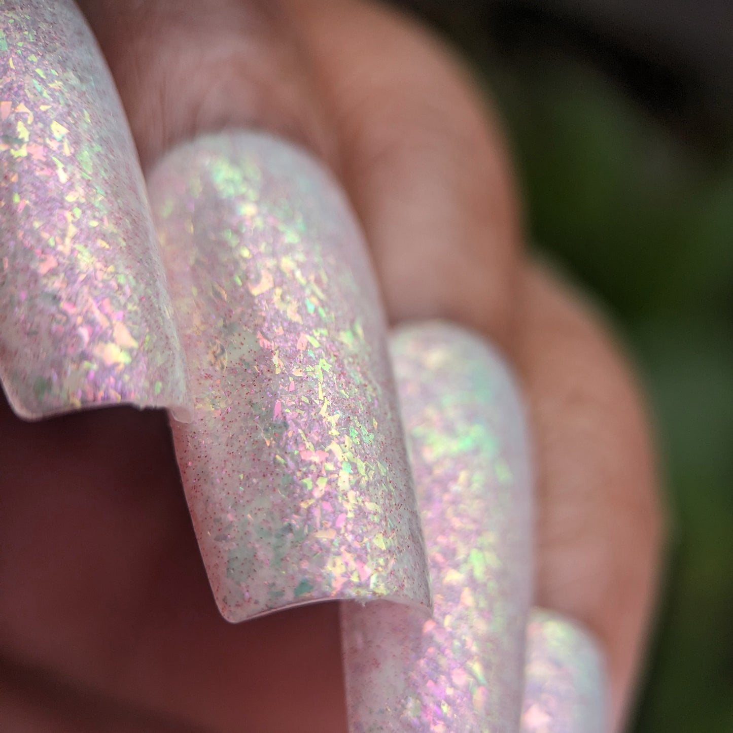 A close-up reveals nails embellished with rainbow flakies and a holographic effect from PI Colors' Caelestis Dreams.005. This handmade polish shimmers in pink, green, and purple under light, while a blurred background enhances the focus on this iridescent masterpiece.
