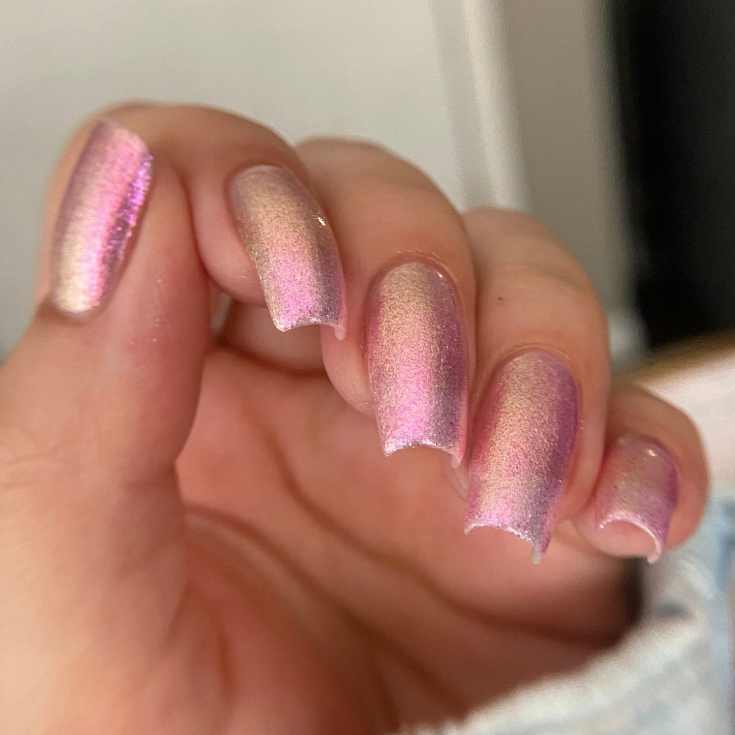 A close-up of a hand with manicured nails painted in a shimmery pink metallic polish, with a light denim sleeve visible at the bottom.