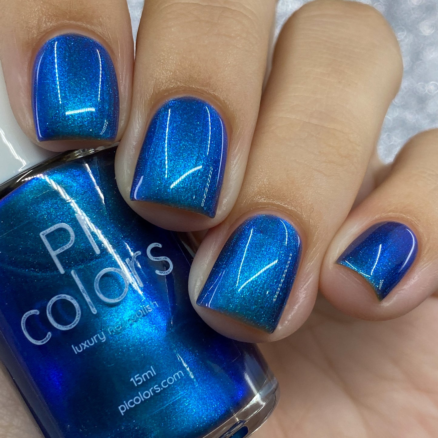 Close-up of a hand adorned with PI Colors' Aurora Blue.316 luxury nail lacquer, its holographic shimmer capturing the light for a radiant finish. The thumb holds a bottle of matching handmade nail polish labeled "Aurora Blue.316.