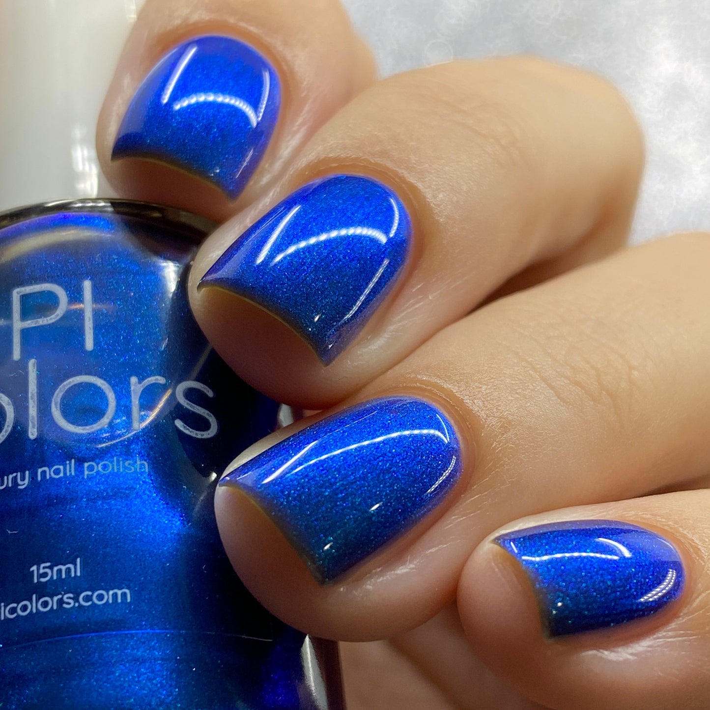 A close-up showcases a hand with short nails painted in Aurora Blue.316, an electric blue nail polish from PI Colors that evokes the vividness of neon signs. In the background, a bottle of PI Colors mirrors this vibrant hue, with both the nails and polish displaying a glossy, reflective finish.