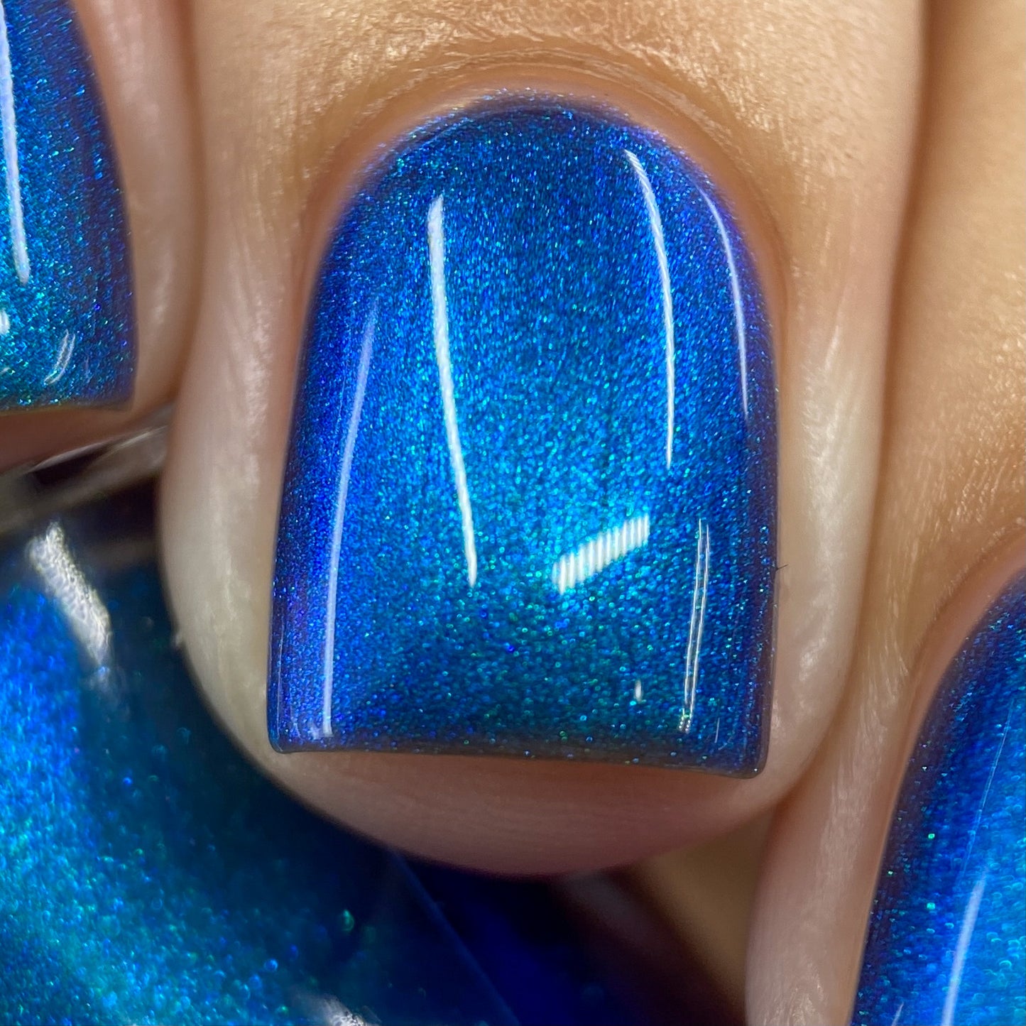 Close-up of a fingernail adorned with PI Colors Aurora Blue.316 luxury nail lacquer. This handmade polish features a glossy, metallic blue hue with a shimmering effect that reflects light, emphasizing its vibrant color. The precise application beautifully highlights the surrounding skin.