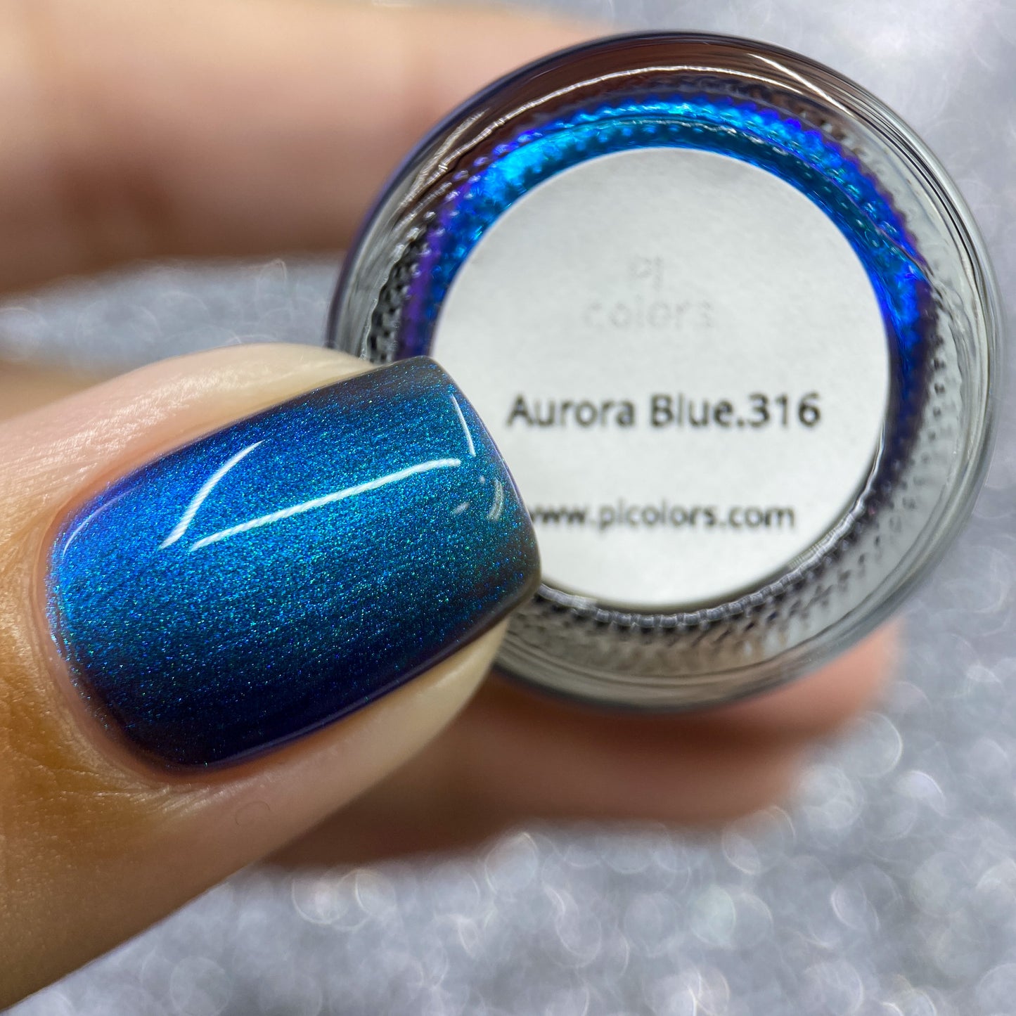 A close-up reveals a fingernail coated in shimmering electric blue nail polish. Nearby, the bottle cap is labeled with "Aurora Blue.316" by PI Colors, highlighting its 10-free formula. The sparkling silver background enhances the elegance of this vibrant display.