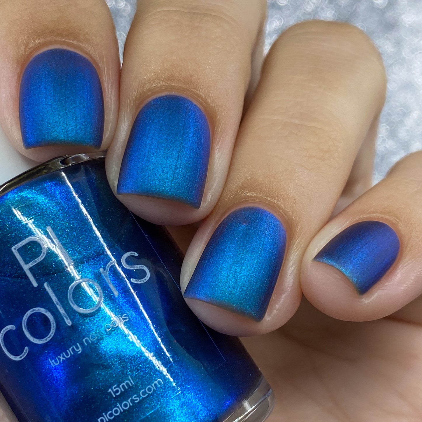 A hand with nails adorned in the glossy Aurora Blue.316 is depicted clutching a bottle of luxury nail lacquer from PI Colors. The label on the bottle matches the vibrant blue polish seen on the nails, while a softly blurred, silver-toned backdrop adds an elegant flair to this artisanal nail polish display.