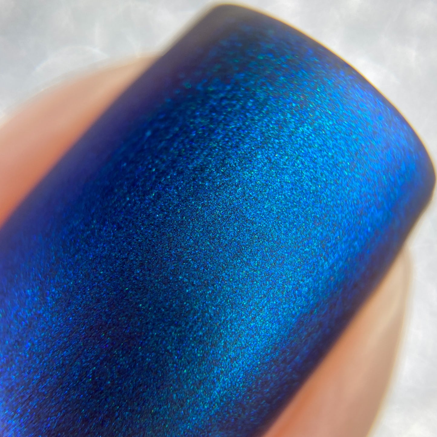A close-up shot of a fingernail showcases the vibrant Aurora Blue.316 nail polish from PI Colors. The 10-free formula delivers a smooth, metallic finish that reflects light with stunning blue gradients. The softly blurred background enhances the texture and brilliance of the nail's appearance.