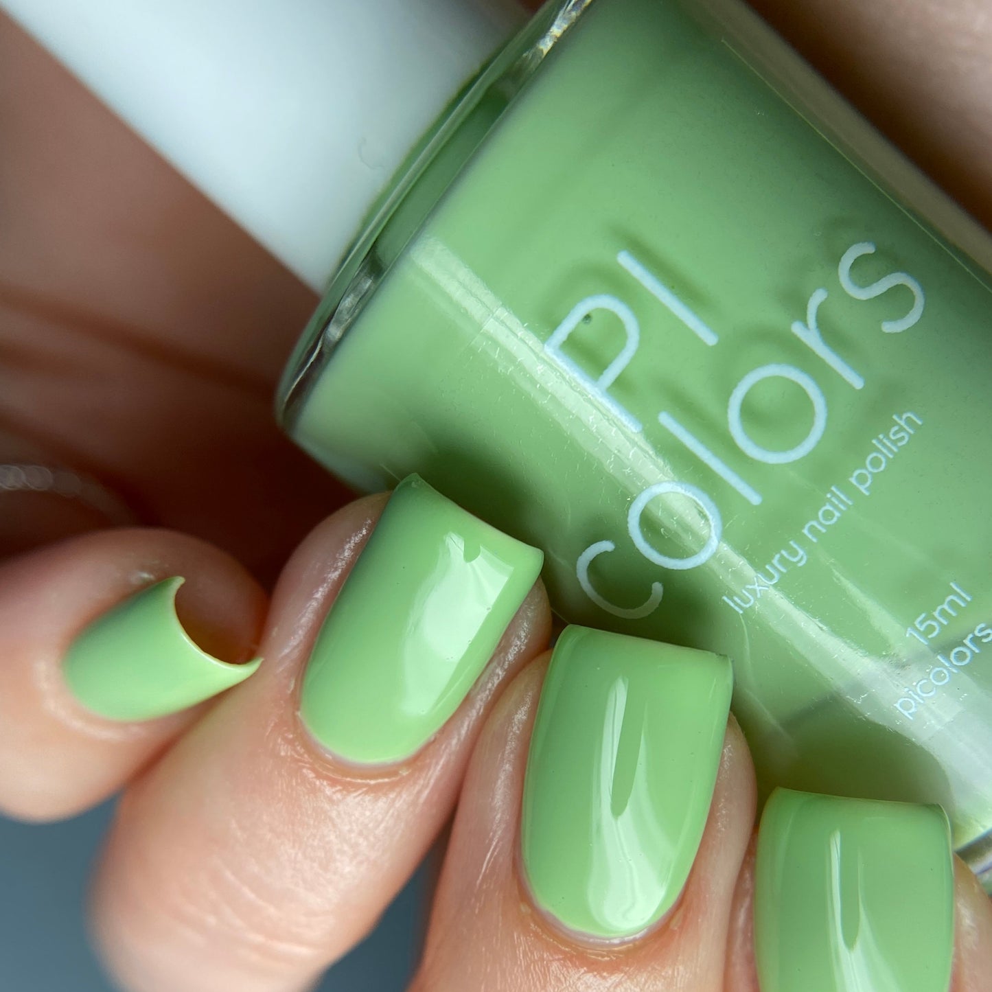 A close-up shows a hand with long, neatly manicured nails painted in a glossy pistachio hue. A bottle of matching creme finish nail polish is held between the fingers, labeled "PI Colors Pistachio.400 Green Nail Polish.