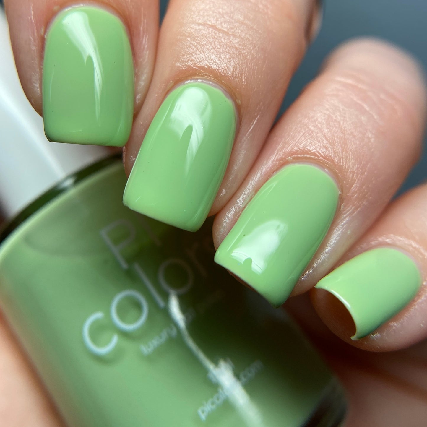A close-up of a hand with nails painted in the glossy shade of PI Colors' Pistachio.400 Green Nail Polish, while a matching bottle of this handmade polish is held slightly blurred in the background.