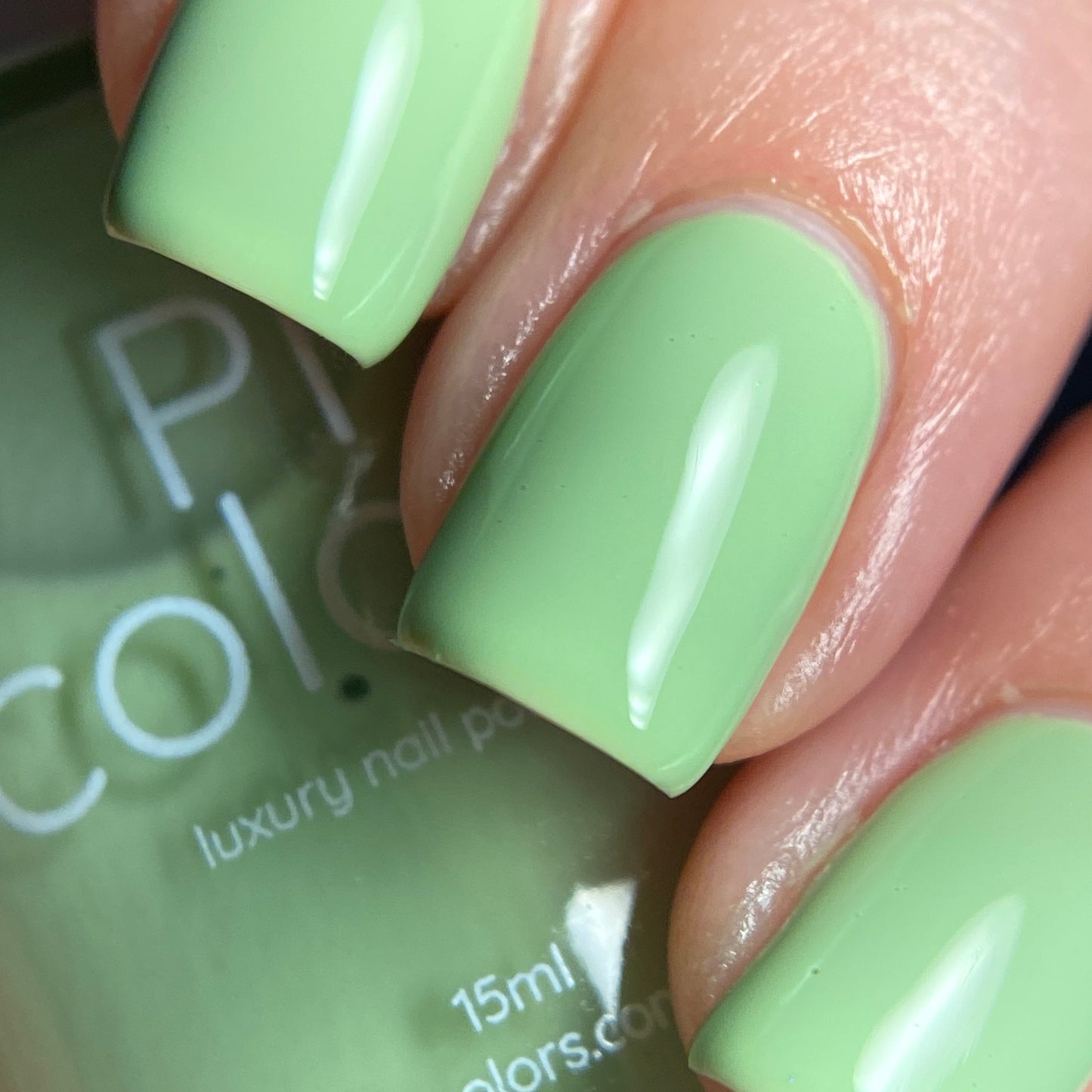 Close-up of fingernails painted with Pistachio.400 Green Nail Polish by PI Colors, showcasing a glossy, pistachio hue with a smooth and shiny finish. The bottle label is partially visible in the background with white text, while the well-manicured nails elegantly highlight the beauty of this creme finish nail polish.