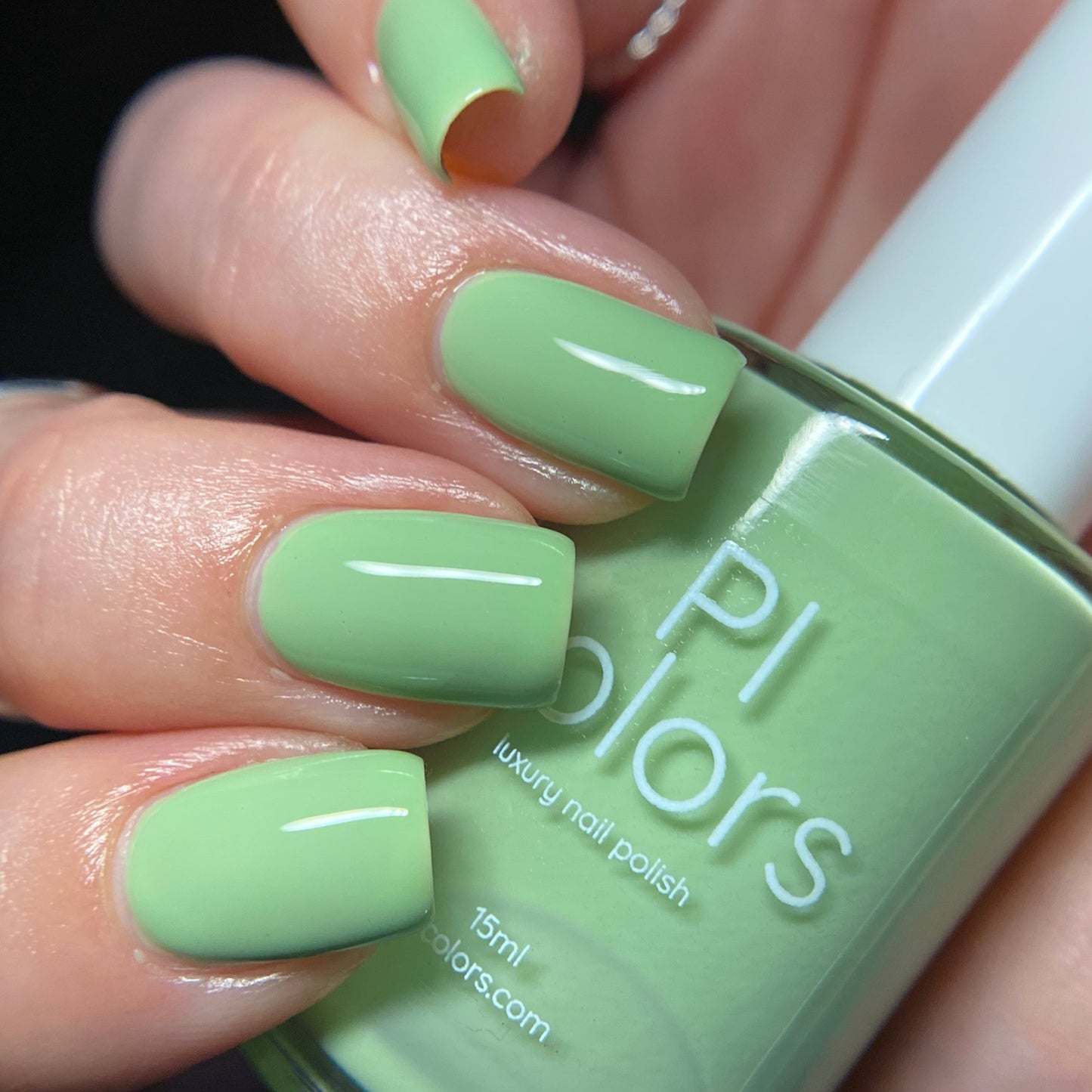 A close-up of a hand adorned with neatly painted light green nails showcases the luxurious Pistachio.400 Green Nail Polish by PI Colors. The creamy finish of the polish is beautifully complemented by the matching bottle, which prominently features the brand name "PI Colors" along with "luxury handmade nail polish" on its label.