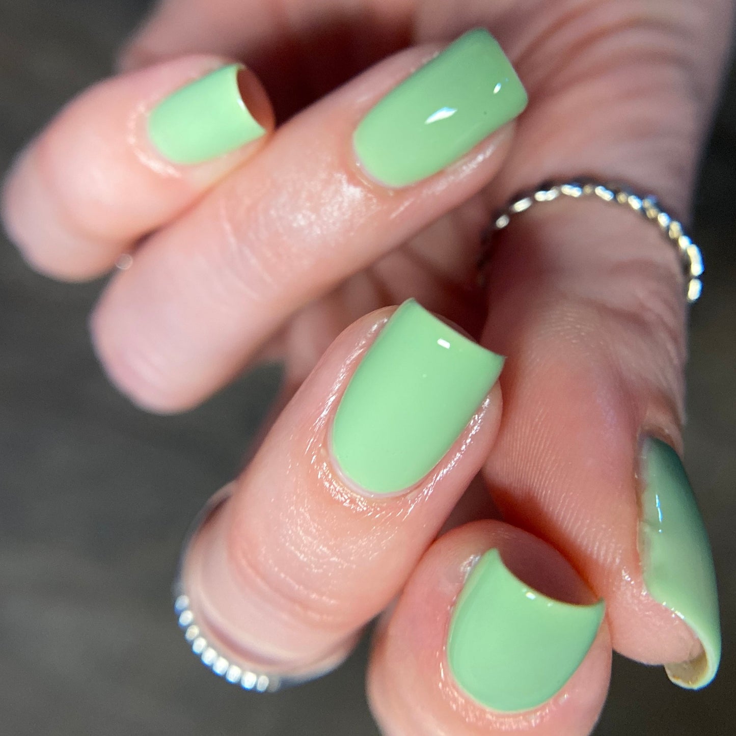 Close-up of a hand with neatly manicured nails painted in PI Colors' Pistachio.400 Green Nail Polish, featuring its vibrant creme finish. The fingers are adorned with thin silver rings, and the background is blurred to emphasize the glossy effect.