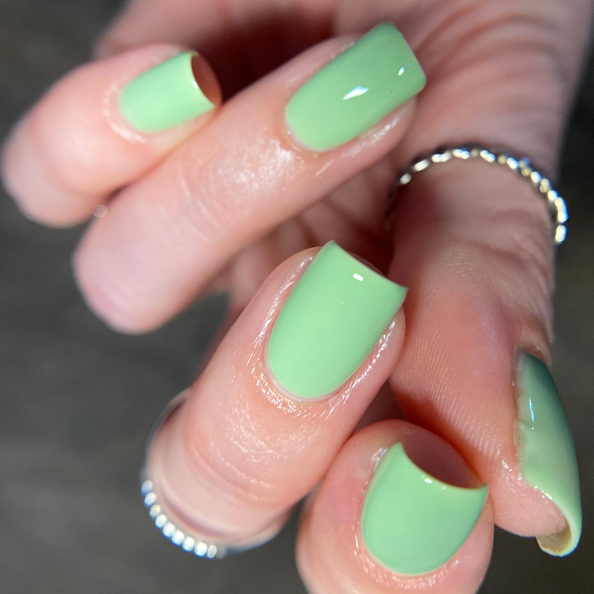 Close-up of a hand with neatly manicured nails painted in PI Colors' Pistachio.400 Green Nail Polish, featuring its vibrant creme finish. The fingers are adorned with thin silver rings, and the background is blurred to emphasize the glossy effect.