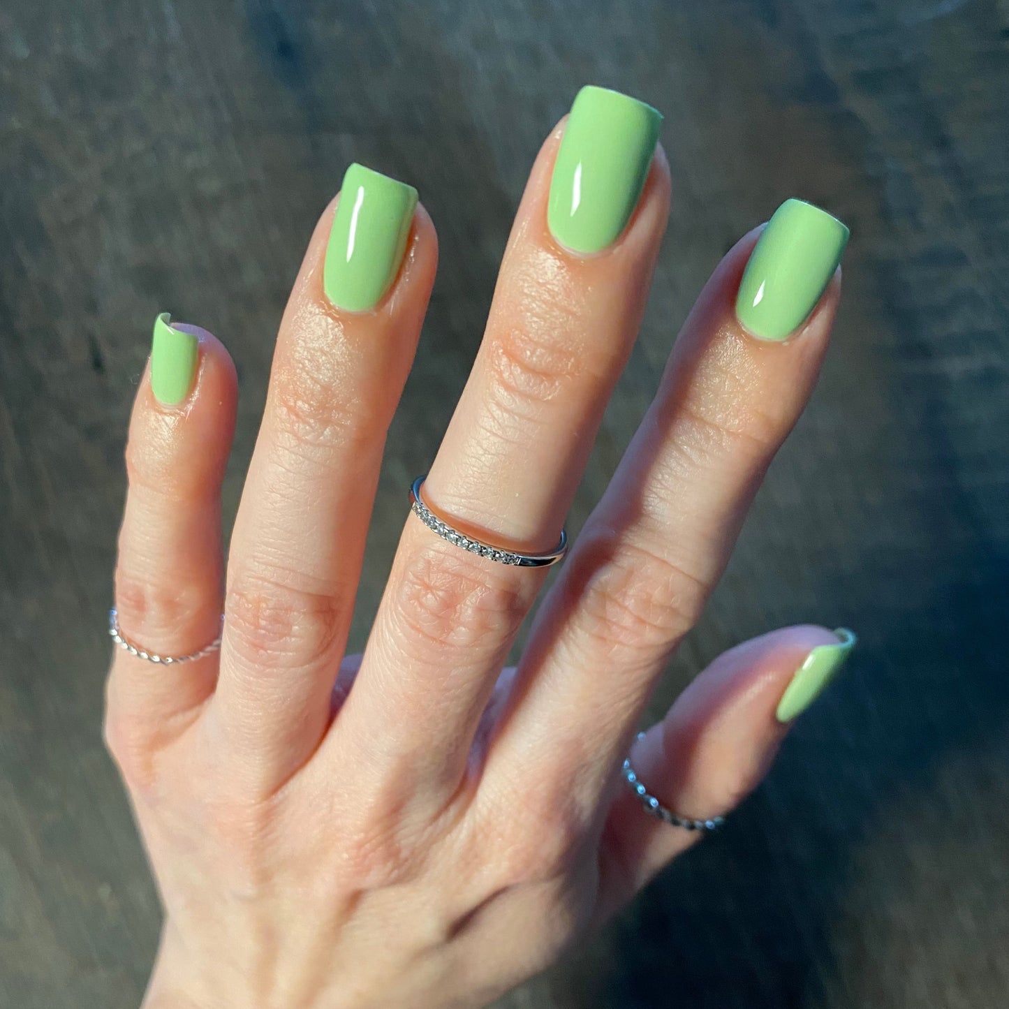 A hand featuring Pistachio.400 Green Nail Polish by PI Colors is displayed on a dark wood surface, with each finger adorned by thin silver rings. The lighting highlights the glossy, crème finish of the nail polish.