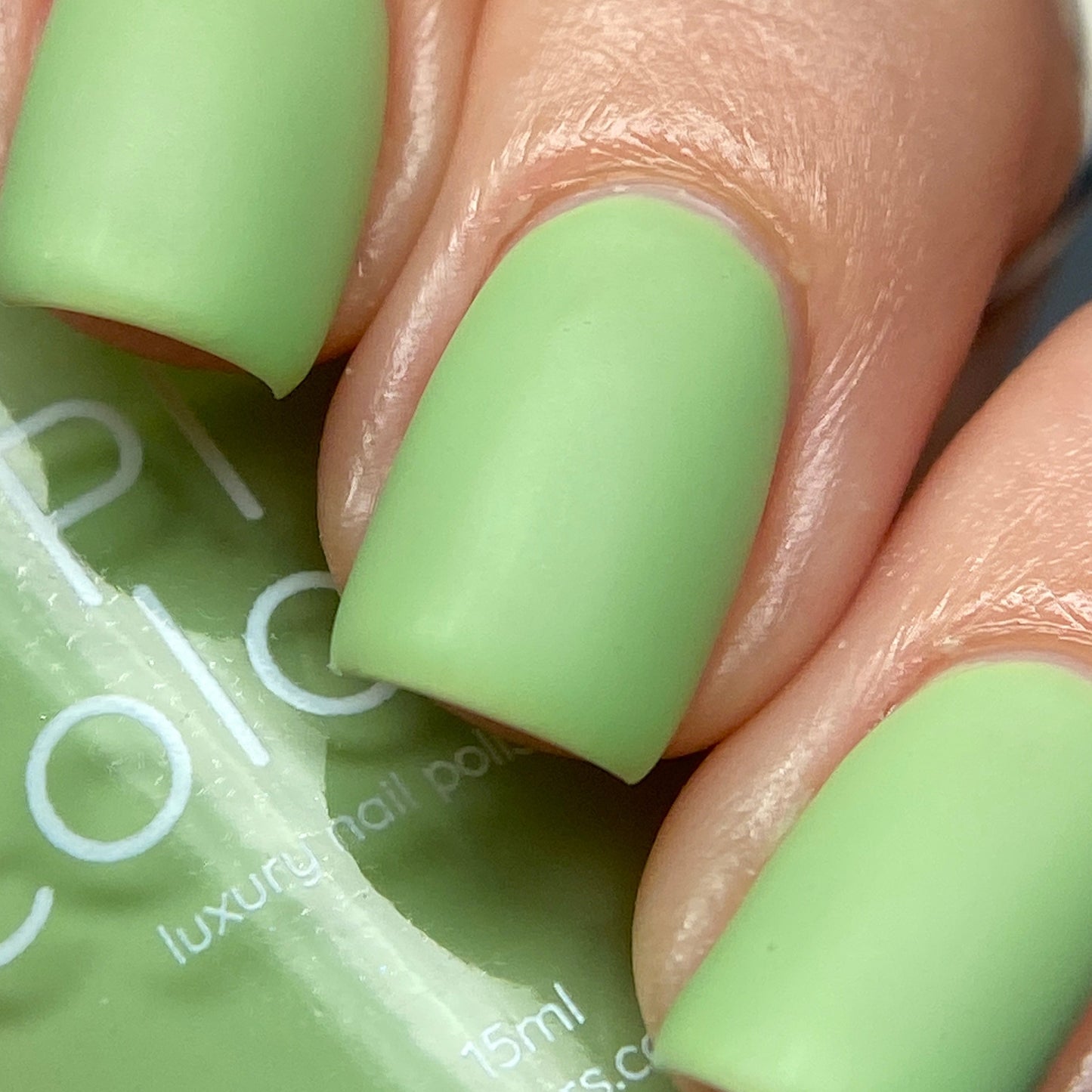 Close-up of a hand with nails painted in a matte pastel pistachio color. The fingers clutch a bottle of matching nail polish labeled "PI Colors Pistachio.400 Green Nail Polish". The nails boast a smooth and even creme finish, showcasing the vibrant, trendy shade typical of luxury nail lacquers.
