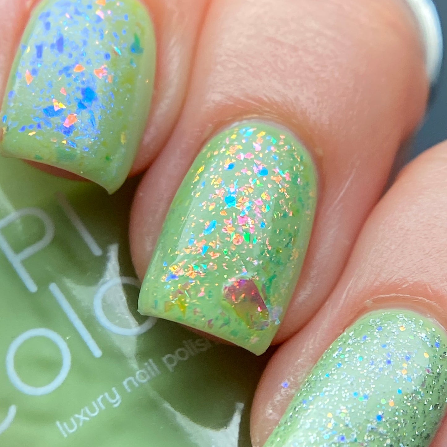 Close-up of fingernails painted with PI Colors' Pistachio.400 Green Nail Polish, showcasing its iridescent, holographic flakes. The shimmering effect creates a colorful pattern on the nails, with a hint of the luxury nail lacquer bottle visible in the background.