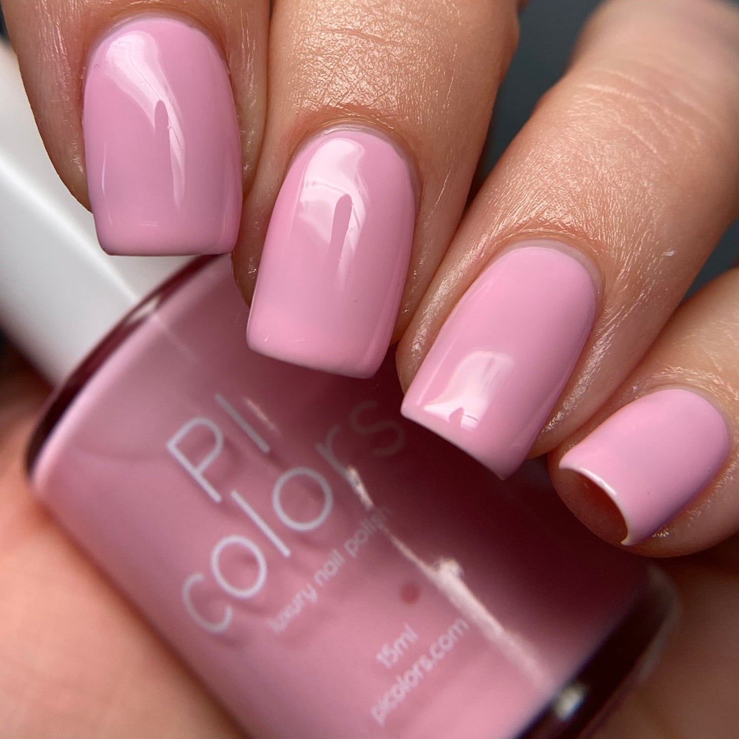 A close-up shot features a hand with nails elegantly painted in a glossy, pastel shade from the Cupcake.400 Pink Nail Polish collection. The fingers cradle a bottle of luxury nail lacquer labeled with the brand "PI Colors," both boasting a sleek and lustrous finish.