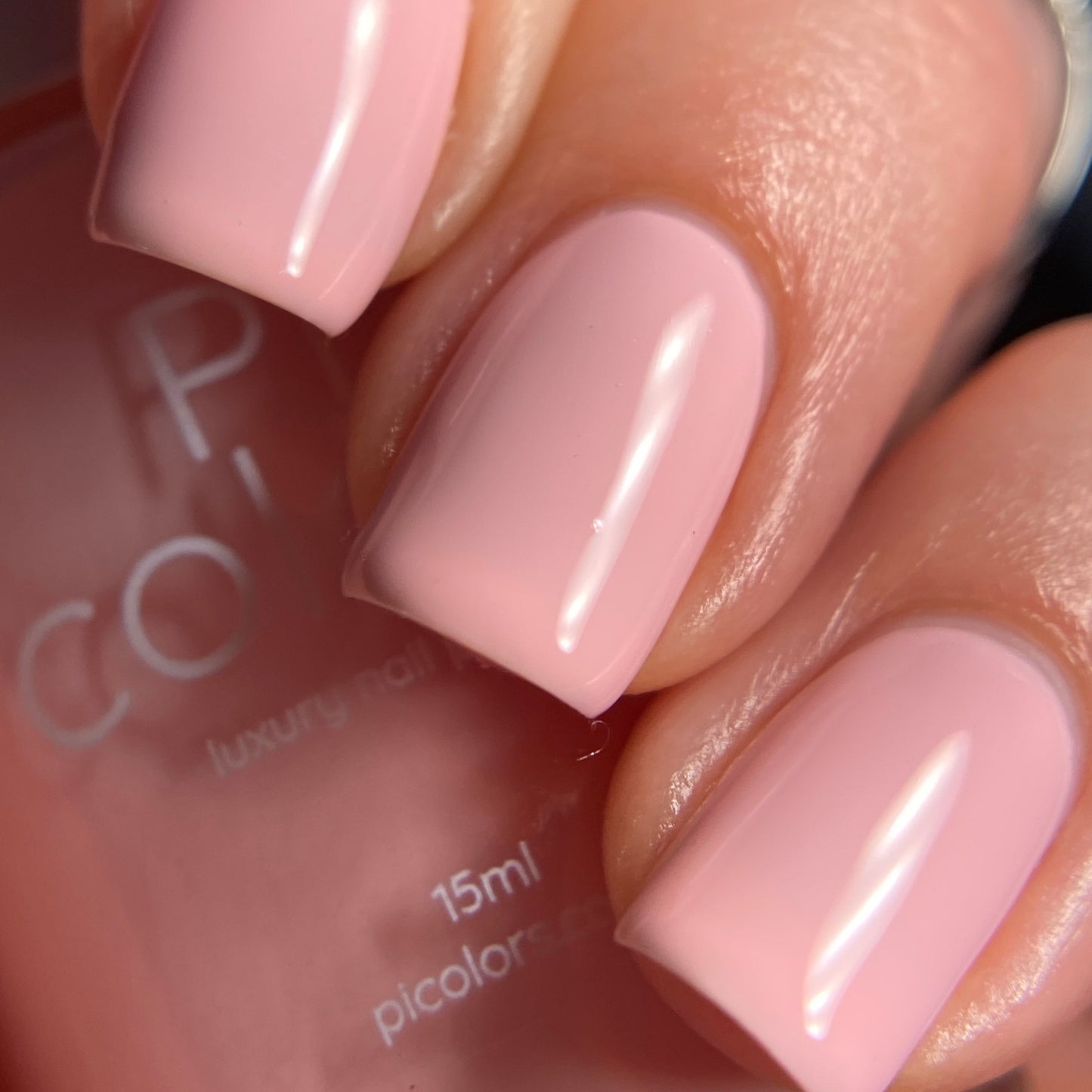 Close-up of a hand showcasing neatly manicured nails painted in a smooth, glossy pastel pink shade with PI Colors' Cupcake.400 Pink Nail Polish. The background includes part of the nail polish bottle with text partially visible.