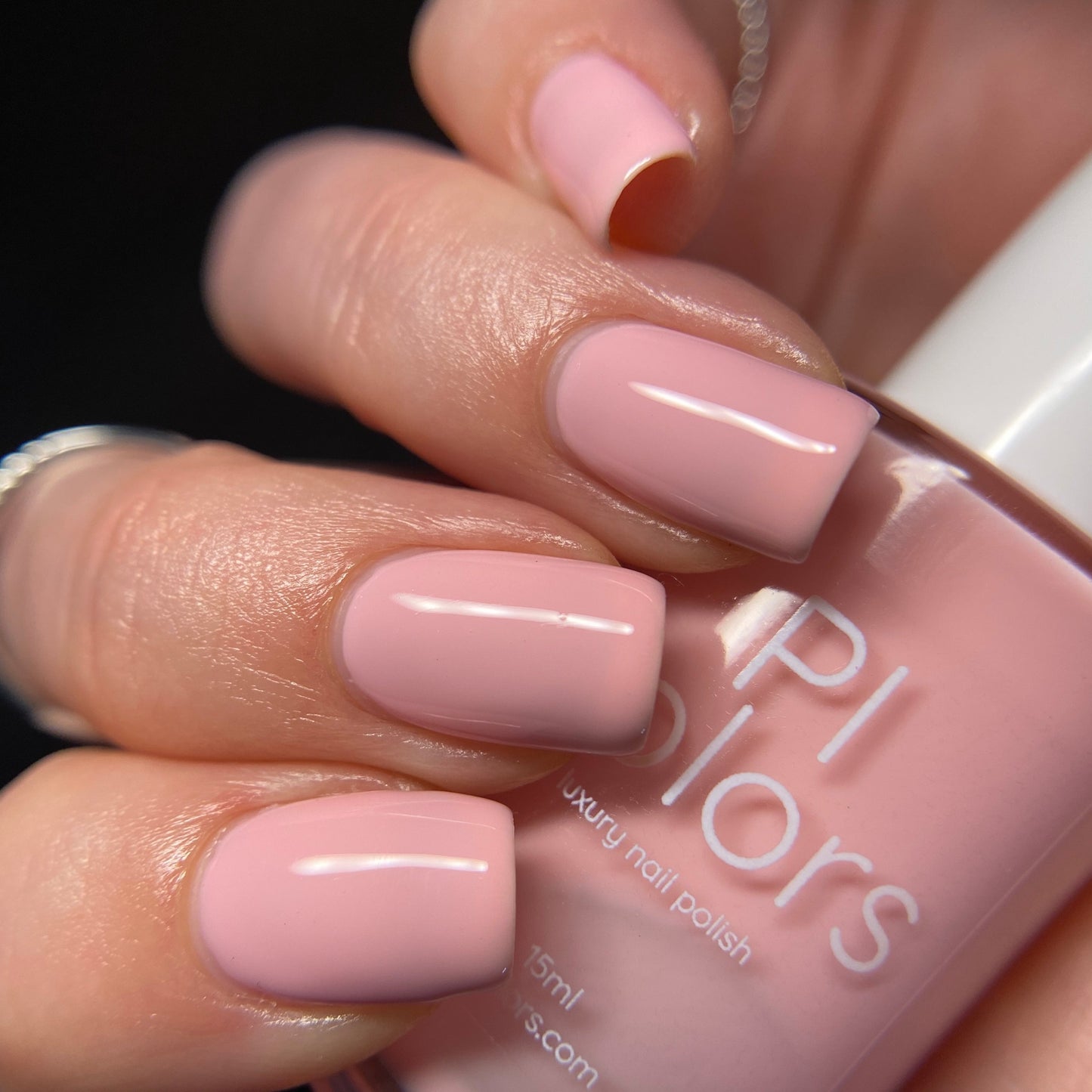 A close-up shot features a hand with neatly manicured fingernails painted in a glossy pastel pink color. The fingers hold a bottle of "Cupcake.400 Pink Nail Polish" from PI Colors. The dark background accentuates the shimmer of the creme finish and highlights the elegant nails beautifully.