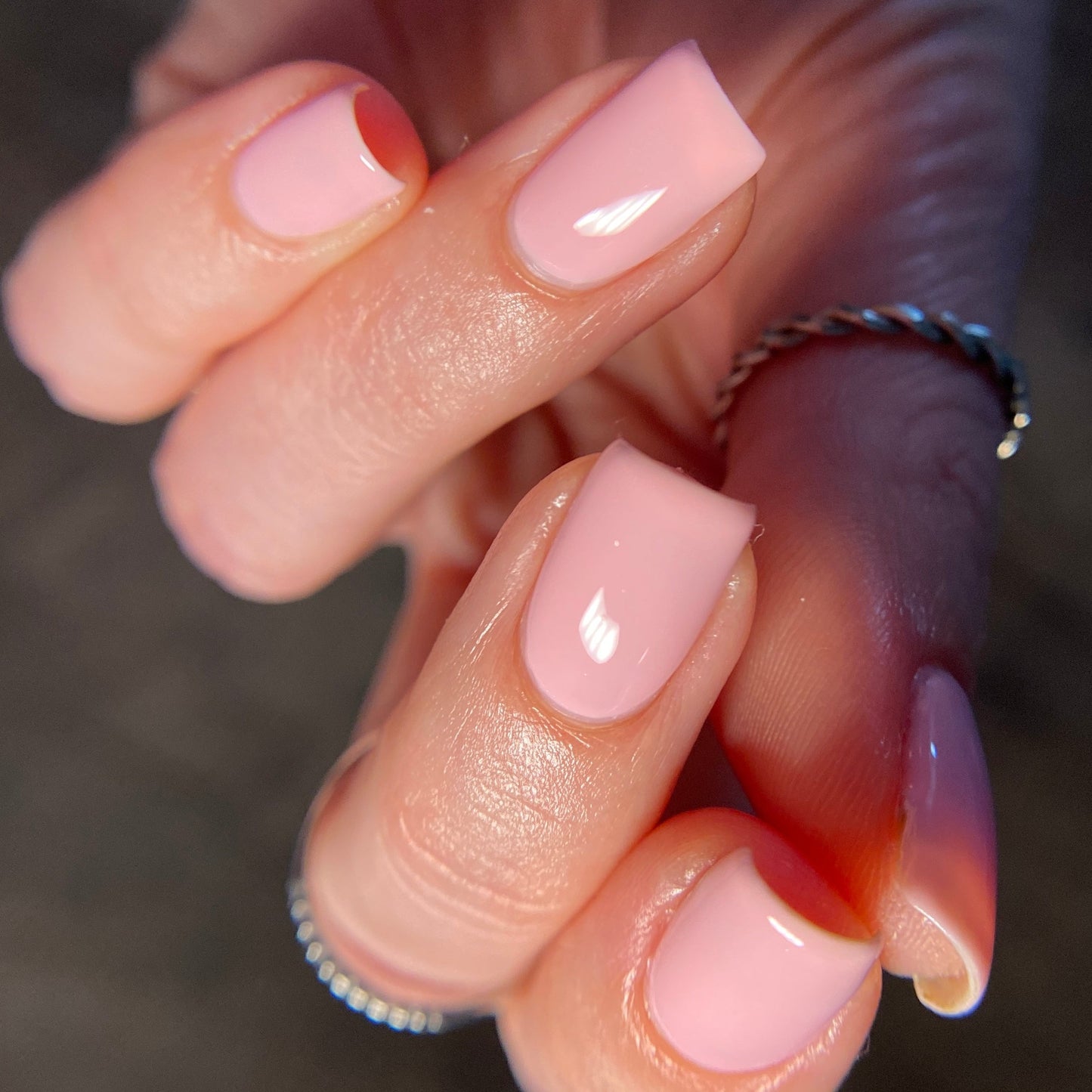 Close-up of a hand displaying neatly manicured nails with PI Colors' luxury Cupcake.400 Pink Nail Polish in pastel pink. The square-shaped nails boast a glossy crème finish, while a delicate silver twisted ring adorns the hand, adding an extra touch of elegance.