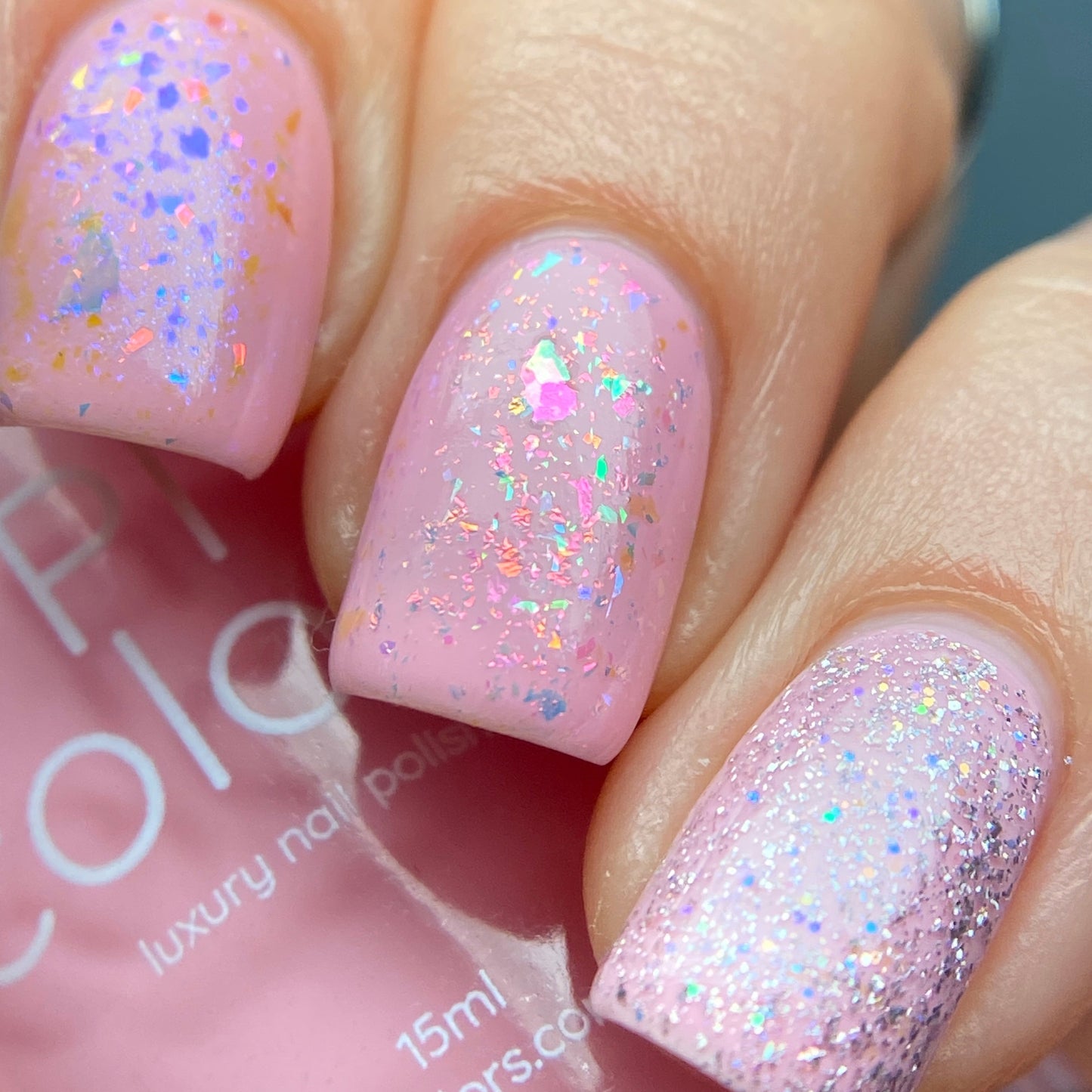 Close-up of fingernails painted with PI Colors' Cupcake.400 Pink Nail Polish in a soft pastel pink hue enriched with iridescent glitter. This 10-free polish boasts a glossy finish with holographic specks, producing a sparkling effect under light. The partially visible bottle is seen in the background.