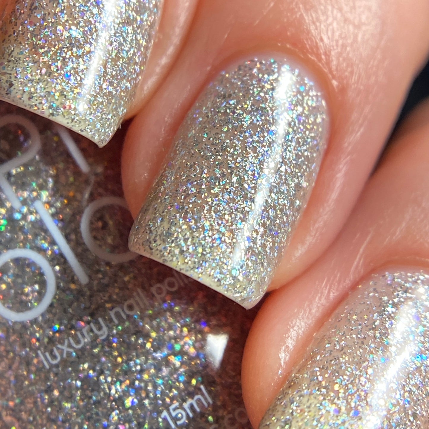 A close-up of a hand reveals nails adorned with PI Colors' Diamond Blast.401 Silver Holographic Glitter Nail Polish Topper, the vibrant hues from the holographic glitter shimmering beautifully in the light. The smooth and glossy finish highlights the brilliance of this luxurious 10-free nail lacquer, with a partially visible bottle elegantly held in the hand.