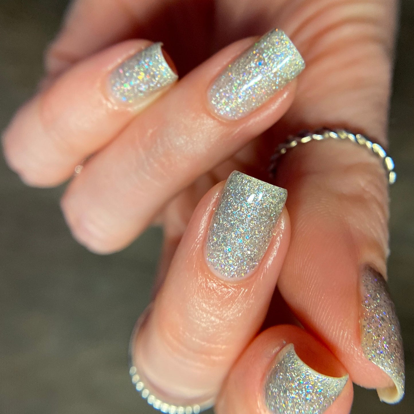Close-up of a hand showcasing PI Colors' Diamond Blast.401 Silver Holographic Glitter Nail Polish Topper on short nails. The hand is embellished with thin silver rings on several fingers, while the blurred background accentuates the sparkle of this luxurious 10-free nail polish.