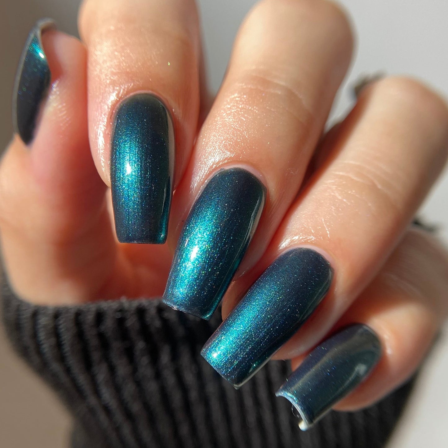 A close-up of a hand with long, almond-shaped nails painted in a metallic, shimmering teal-blue nail polish, wearing a dark ribbed knit sleeve.