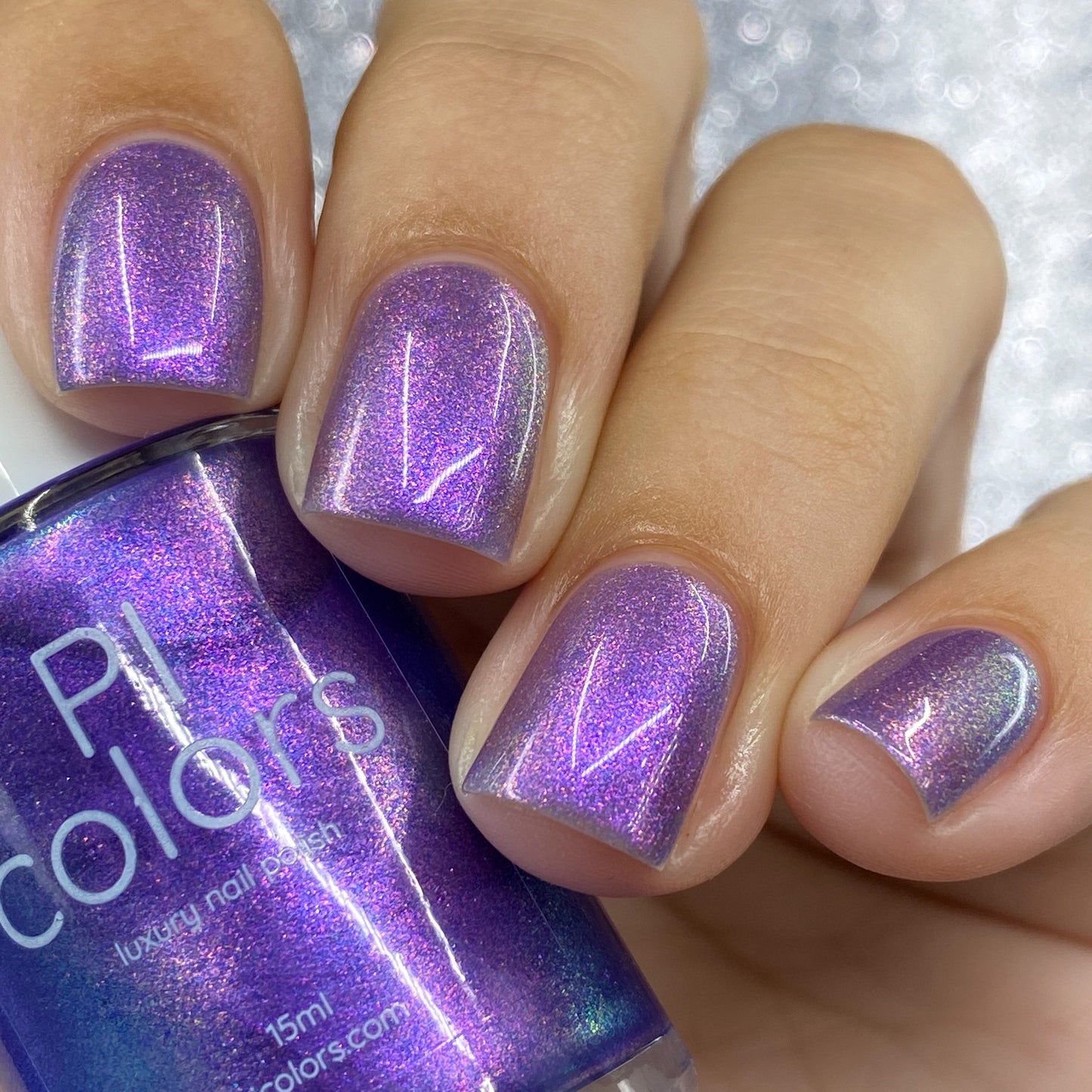 Close-up of fingernails painted with Dusk Rainbow.321 Purple Nail Polish, showcasing its aurora shimmer. The nail polish bottle is held in hand, prominently displaying the PI Colors label against a sparkly, textured backdrop.