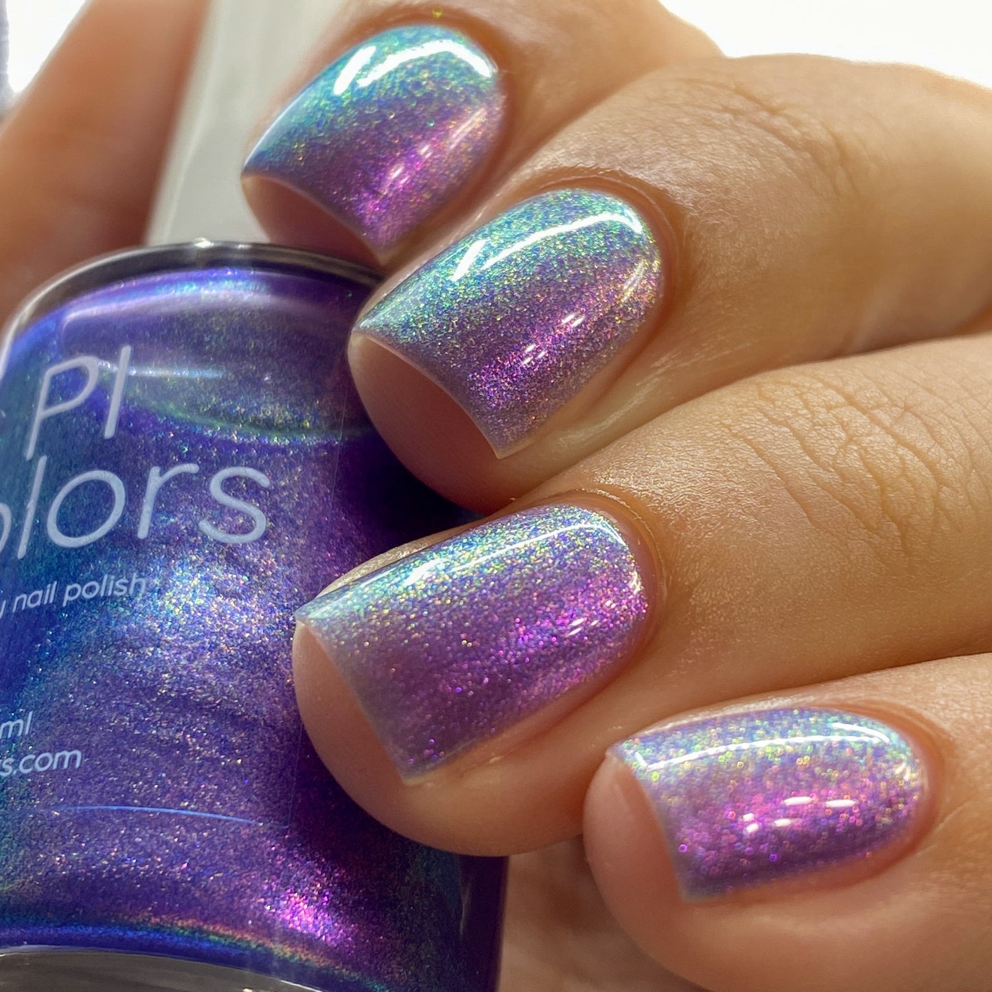 A close-up of a hand showcases nails painted in Dusk Rainbow.321 by PI Colors, a deep purple nail polish with cyan and green aurora shimmer. The same handmade nail polish bottle is held, with its label partially visible. The nails exhibit a glossy, iridescent finish that shifts with the light.
