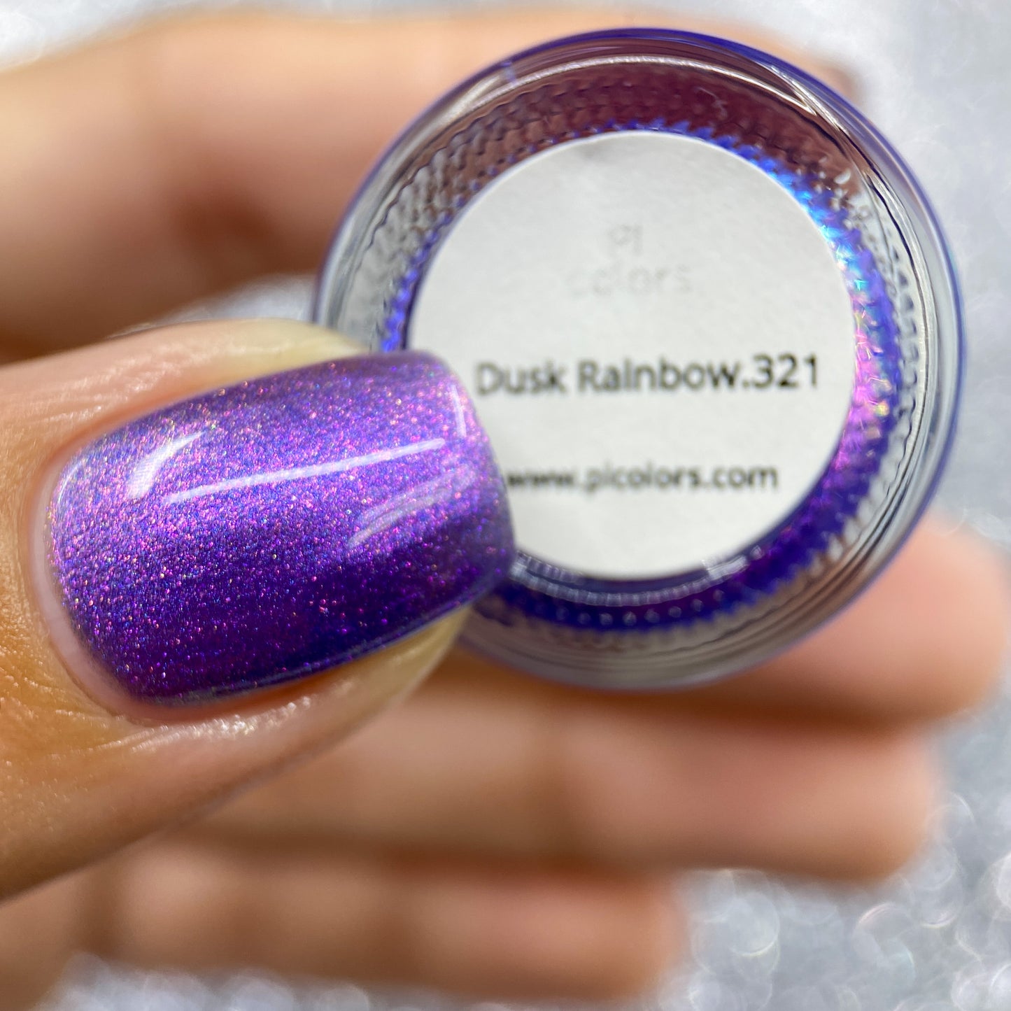 A hand is shown holding a compact container labeled "Dusk Rainbow.321 Purple Nail Polish" by PI Colors, alongside a website URL. The person's fingernail is adorned with the same shimmering deep purple polish as the product. The background features a sparkly silver texture, capturing the charm of this handmade nail polish.