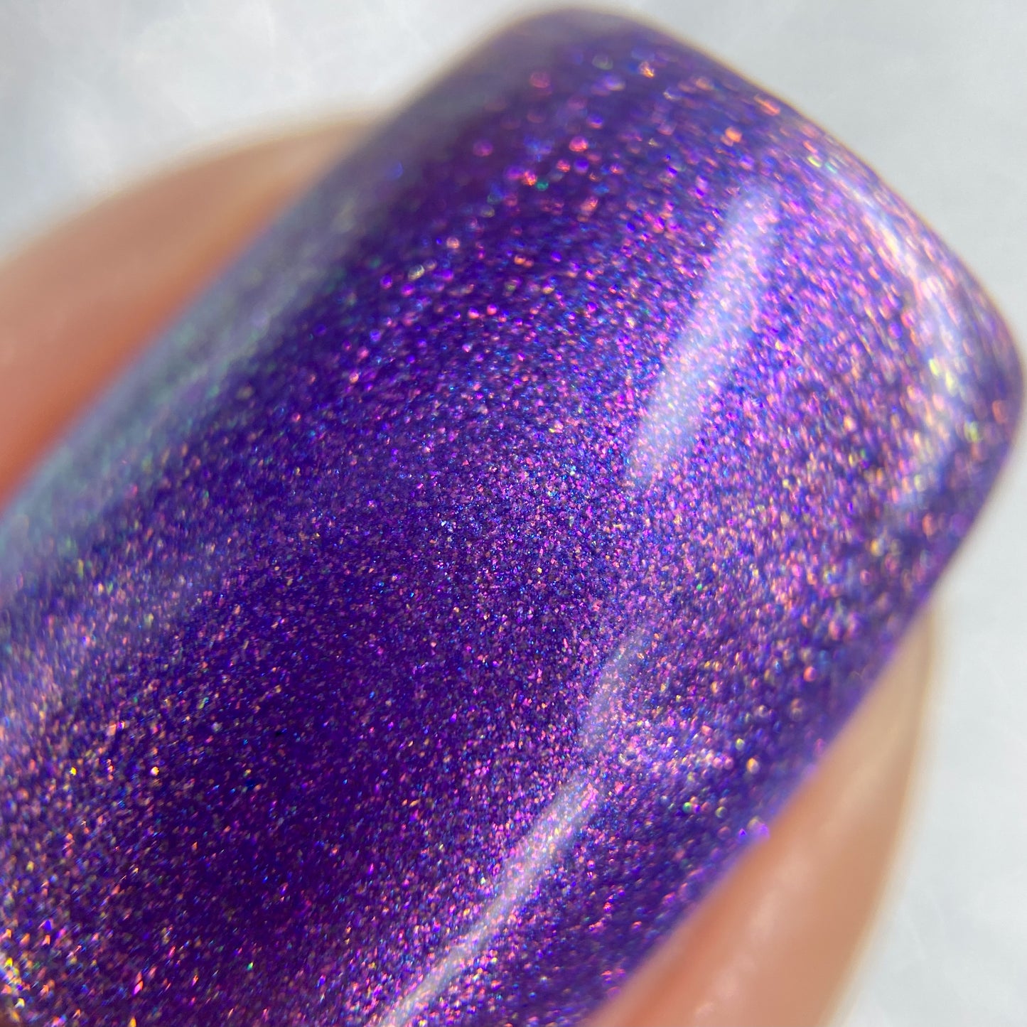 A close-up of a fingernail adorned with PI Colors' Dusk Rainbow.321 Purple Nail Polish reveals a deep purple hue enhanced by a shimmering cyan and green aurora, showcasing a handmade glittery, iridescent texture that dances elegantly under the light.