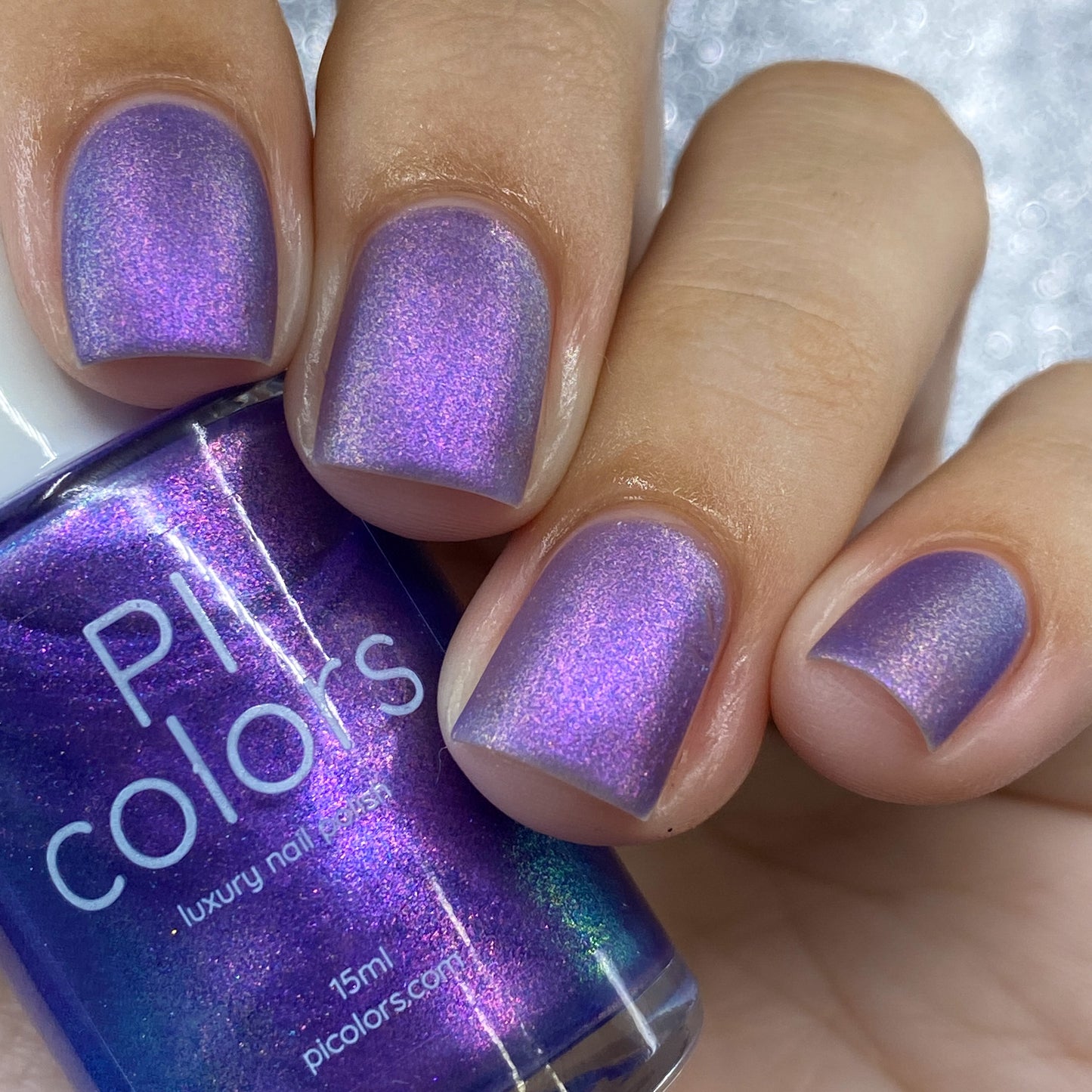 A close-up captures a hand with nails adorned in "Dusk Rainbow.321 Purple Nail Polish" by PI Colors, revealing an alluring cyan and green aurora shimmer. The bottle, which the hand holds, is labeled "PI Colors" and perfectly mirrors the captivating iridescence that shifts from blue to pink under the light.