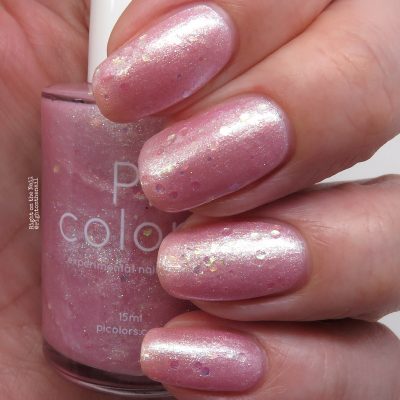 Close-up of fingers painted with shimmery pink nail polish, showcasing PI Colors Pink Candy.210 Nail Polish. The nails have a glossy finish, while the bottle gleams in the background. This pastel pink jelly polish features a pearlescent look with subtle glitter accents.