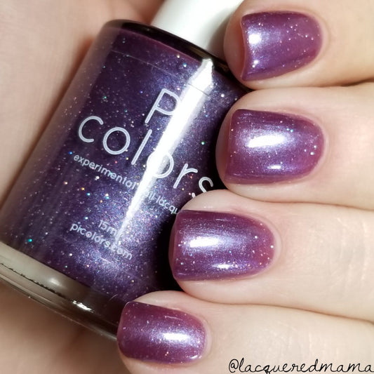 A hand with nails painted in the breathtaking shade named "Blackberry Dreams" holds a bottle of the same hue. The PI Colors bottle features this 10-free polish, enhanced with shimmery rainbow glitter that delivers captivating, sparkly accents.