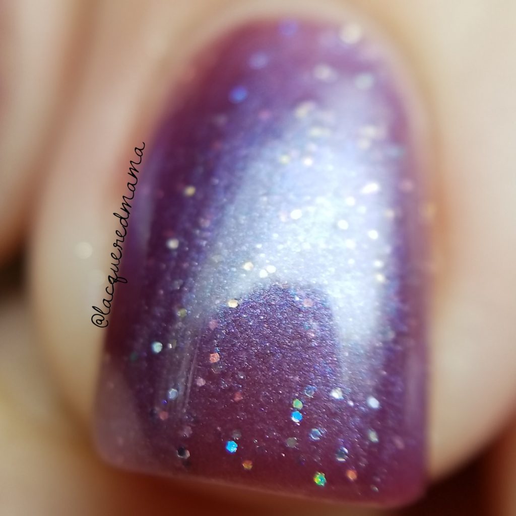 Close-up of a fingernail adorned with PI Colors' Blackberry Dreams.015 Red Purple Nail Polish, showcasing its glossy finish accentuated by shimmering rainbow glitter particles for a dazzling effect. The blurred background emphasizes the intricate details and the brilliance of its 10-free formula.
