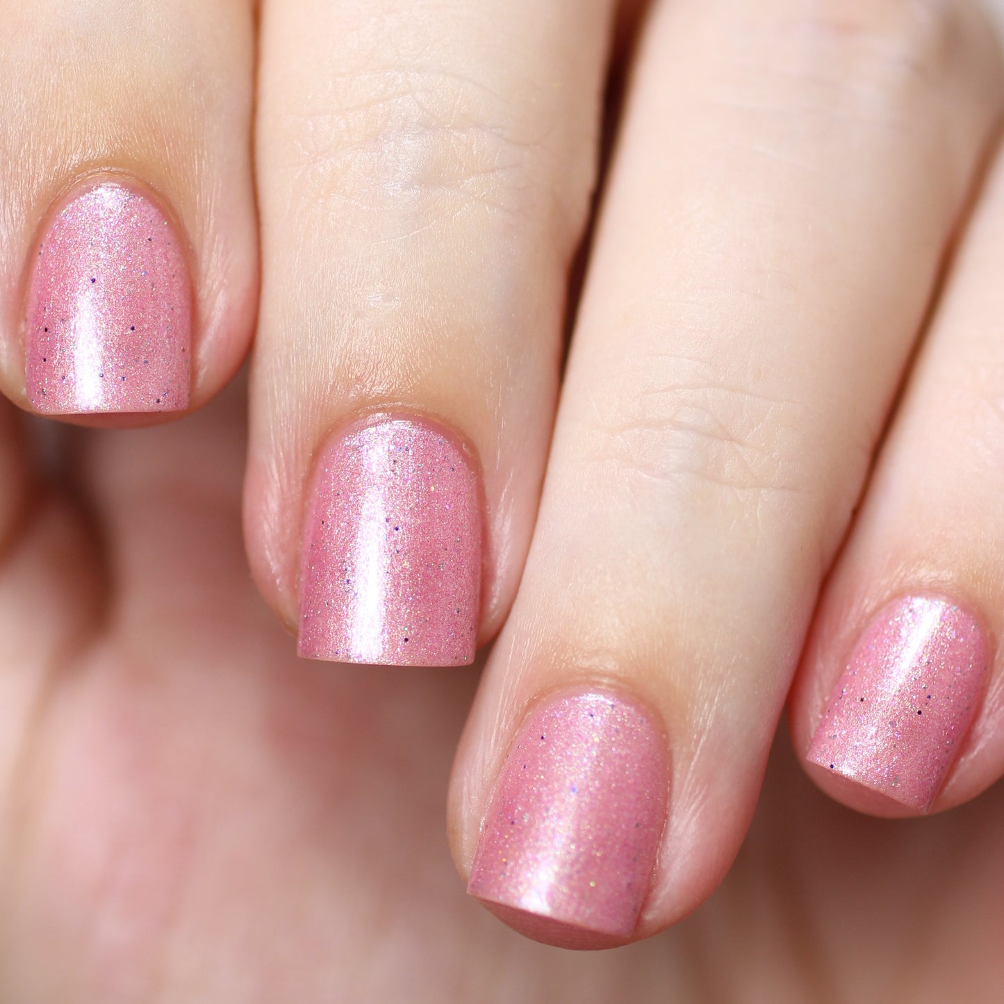 Close-up of a hand with neatly manicured nails painted in PI Colors' Roseate Bliss.012 Pink Nail Polish, featuring a shimmering pink hue. The nails gleam with holographic glitters and have a glossy finish. The smooth, well-cared-for skin beautifully complements the vibrant look of this 10-free polish.