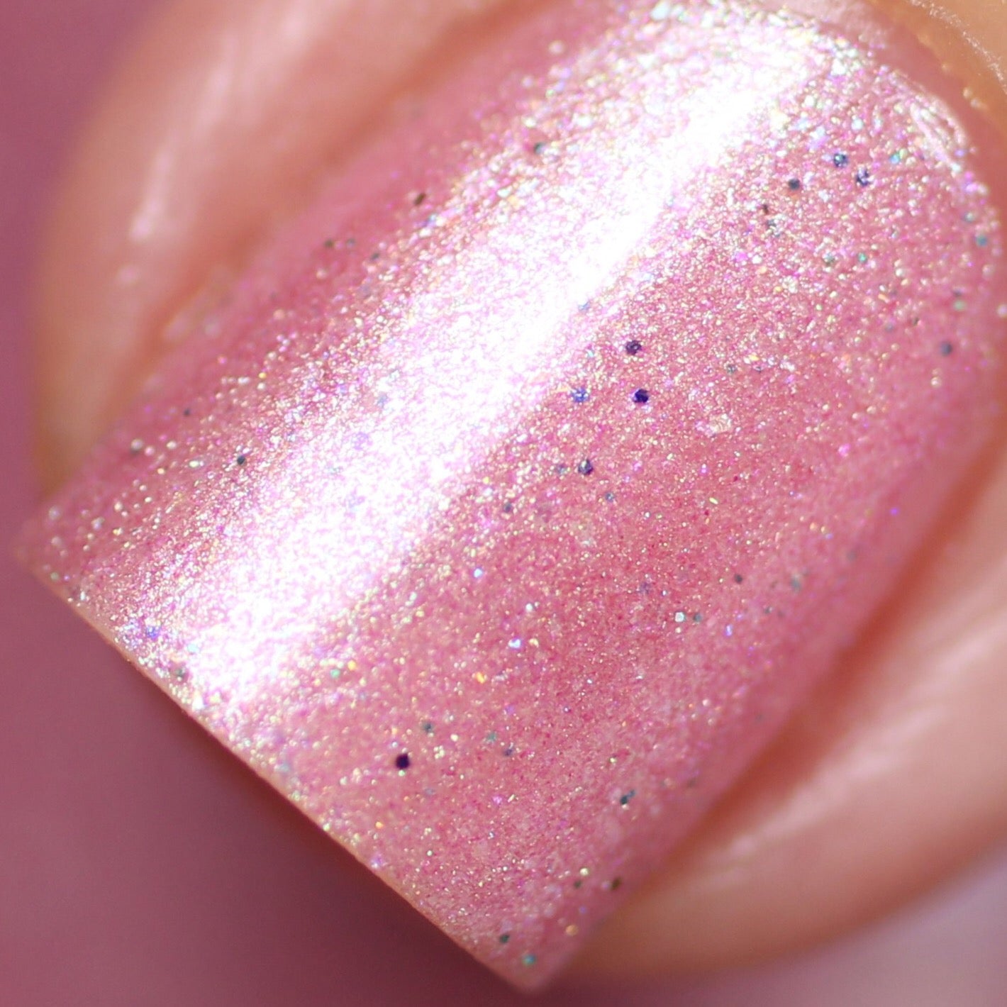 A close-up of a fingernail painted with Roseate Bliss.012 Pink Nail Polish by PI Colors, showcasing its shimmering pink hue adorned with tiny specks and holographic glitters. This 10-free nail polish provides a glossy, iridescent finish that elegantly reflects light, highlighting the nail's smooth and flawless surface.