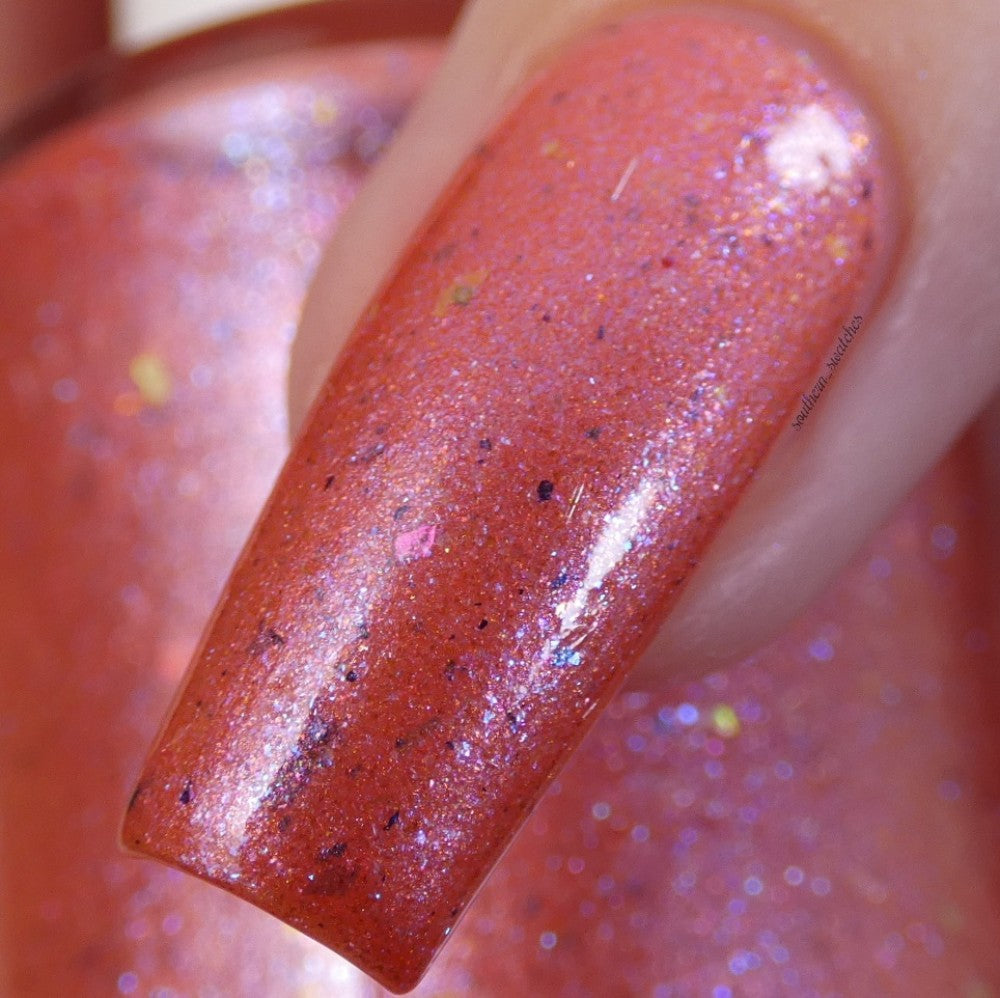 A close-up of a fingernail adorned with Lala Ngoxolo Kumkani Coral Nail Polish by PI Colors, featuring shimmery red tones infused with iridescent rainbow flakes that showcase flecks of pink, purple, and yellow. The glossy finish adds a sparkling allure.