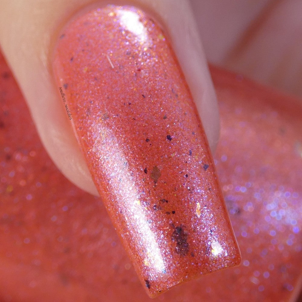 Close-up of a fingernail adorned with PI Colors' Lala Ngoxolo Kumkani Coral Nail Polish, showcasing a shimmering coral pink hue accentuated by reflective glitter particles and iridescent rainbow flakes. The glossy finish gives it a smooth texture, with tiny sparkles visible throughout.