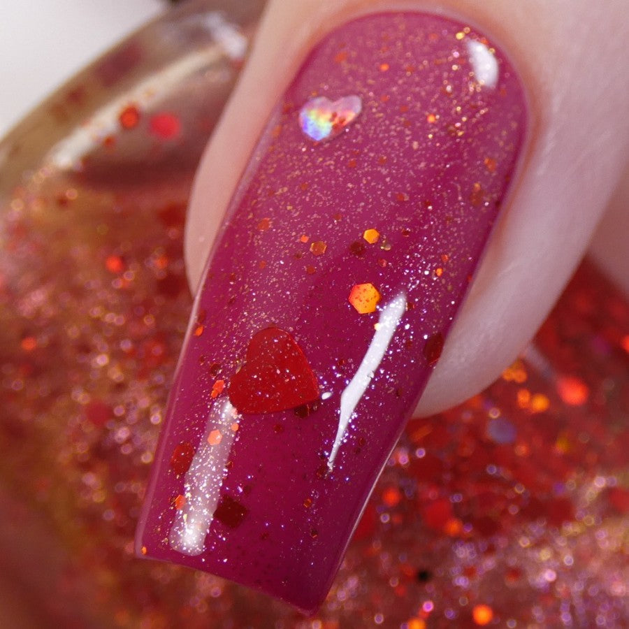 A close-up view showcases a fingernail adorned with PI Colors' Strawberry Heart.208 Red Nail Polish Topper, sparkling with holographic heart-shaped glitter against a pink backdrop. In the blurred background, a bottle of 10-free matching nail polish completes the scene.