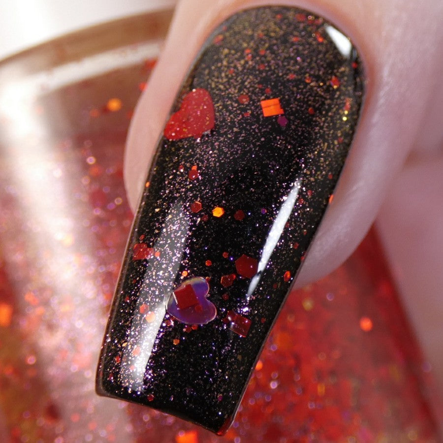 A close-up of a fingernail showcasing the black, 10-free PI Colors Strawberry Heart.208 Red Nail Polish Topper glistening with glitter and holographic heart-shaped confetti catches the eye. A blurred red backdrop enhances this striking nail art.