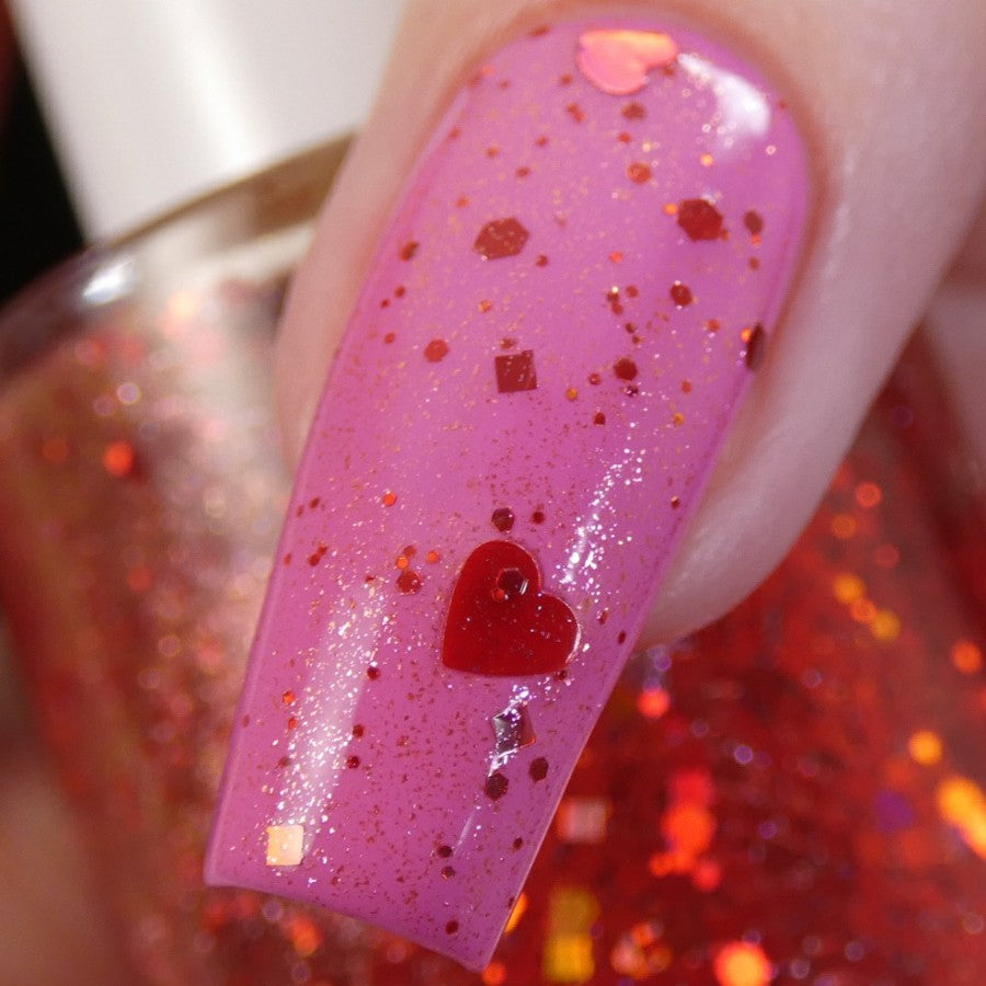 Close-up of a fingernail painted with pink nail polish, adorned with red glitter and holographic heart-shaped sequins. A 10-free bottle of PI Colors' Strawberry Heart.208 Red Nail Polish Topper sits in the background, catching the light.