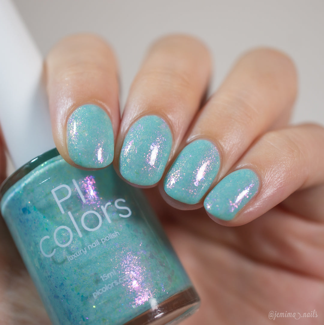 A hand displays pastel teal nail polish with a shimmery, iridescent finish. Colorshifting rainbow flakies glimmer as a bottle labeled "Star of Siam.000 by PI Colors" is held between the fingers, perfectly matching the nails. The softly blurred background emphasizes the glossy texture of the nails.
