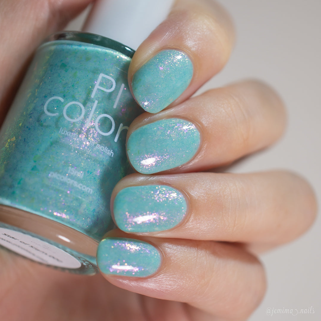 A hand holds a bottle of Star of Siam.000 nail polish by PI Colors. The nails are painted in shimmering turquoise hues with an iridescent finish. The label on this 10-free nail polish is partially visible, promising both vibrant color and a commitment to safe beauty standards.