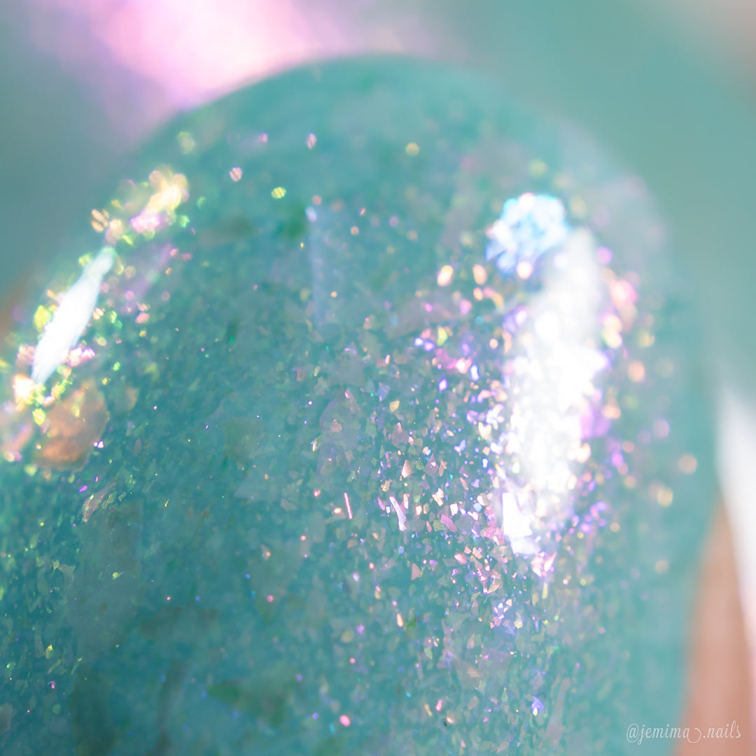 Close-up of a fingernail adorned with PI Colors' Star of Siam.000, an iridescent turquoise nail polish that showcases color-shifting rainbow flakies. This 10-free nail polish glimmers with captivating hues of pink, purple, and blue, resulting in a stunning glass-like effect.